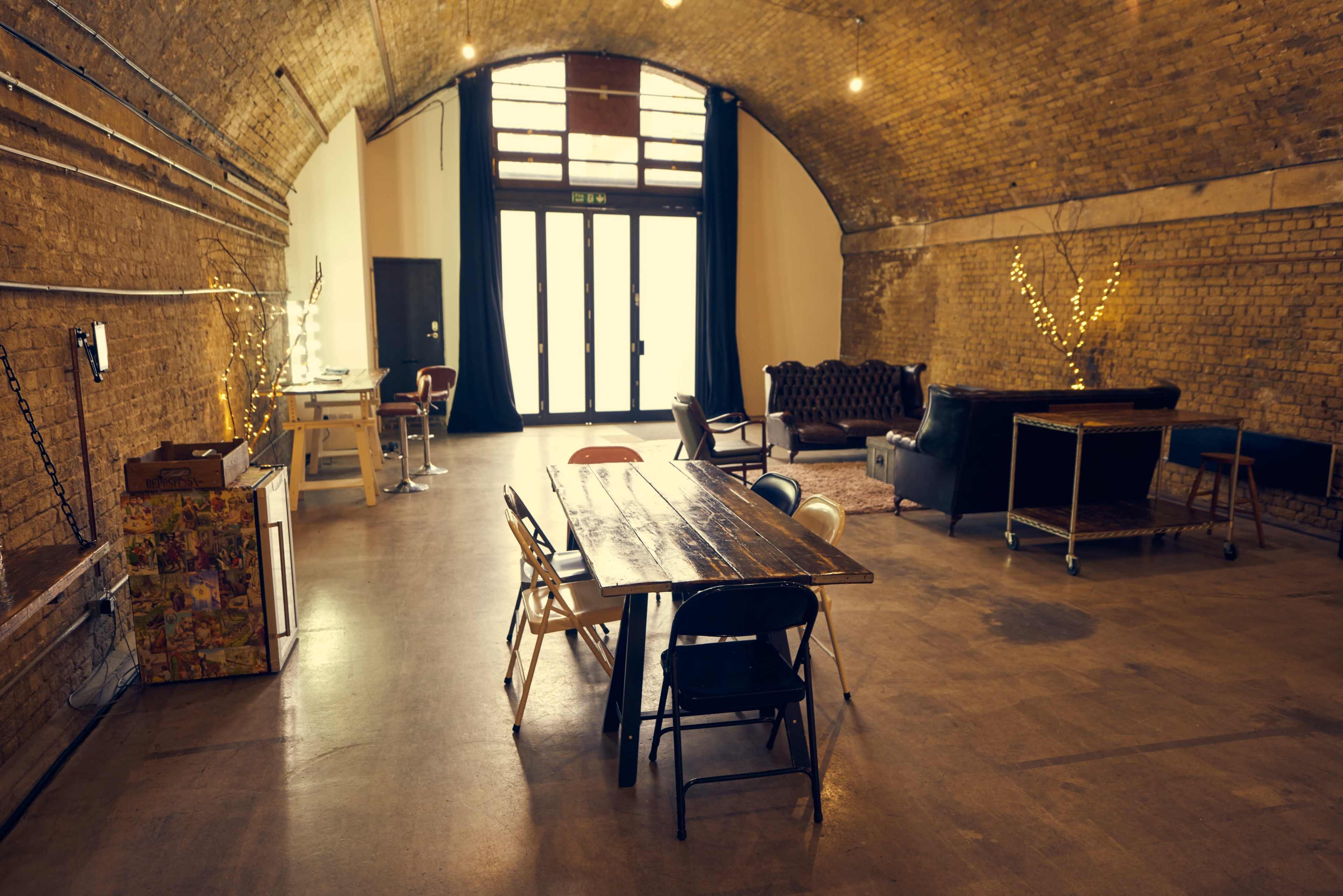 The image shows a spacious room with high arched ceilings, featuring various seating arrangements, a large wooden table, and exposed brick walls.