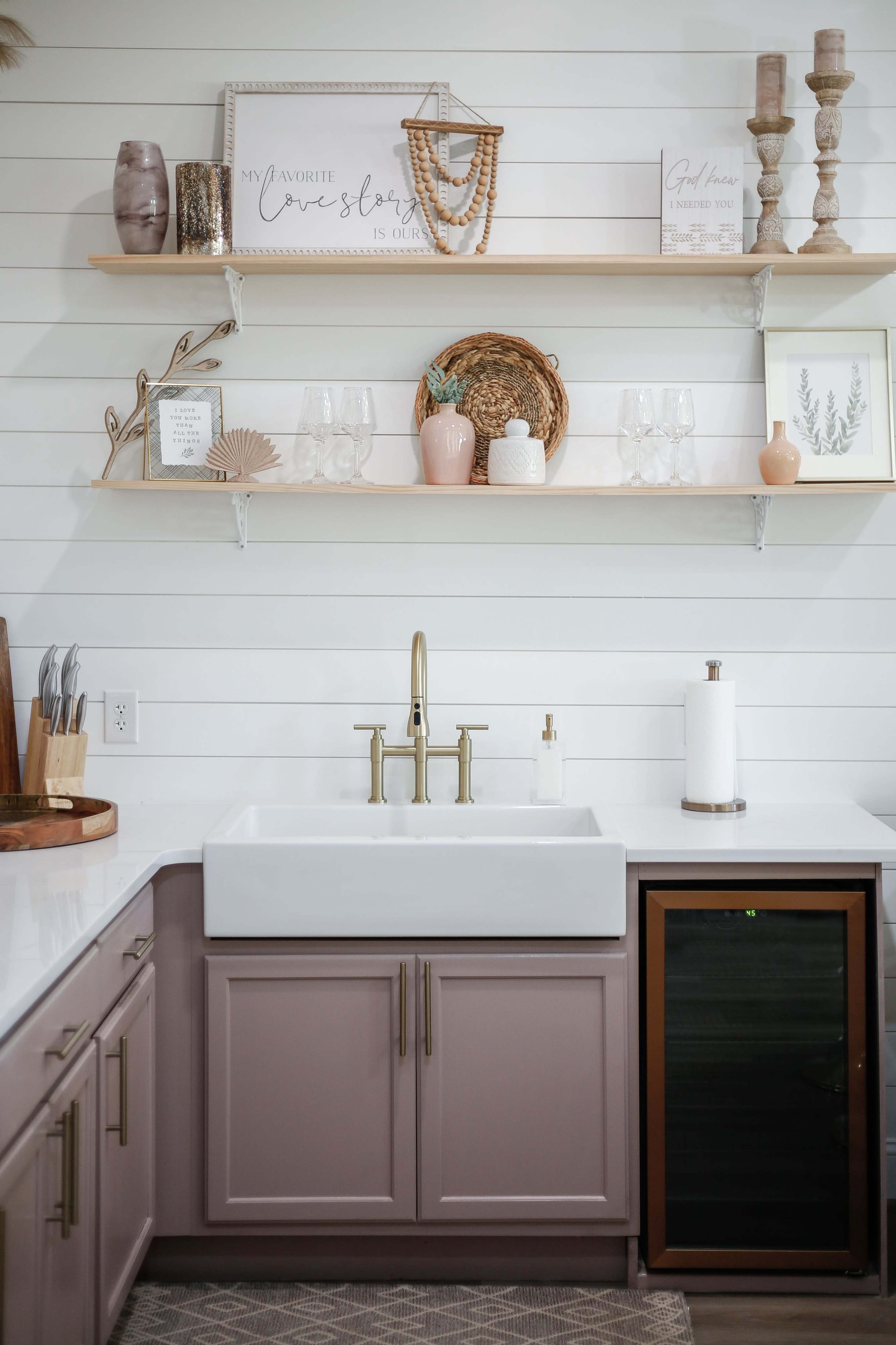 A kitchen sink with a white farmhouse basin, gold faucet, and storage cabinets below, accompanied by decorative shelves above displaying various items.