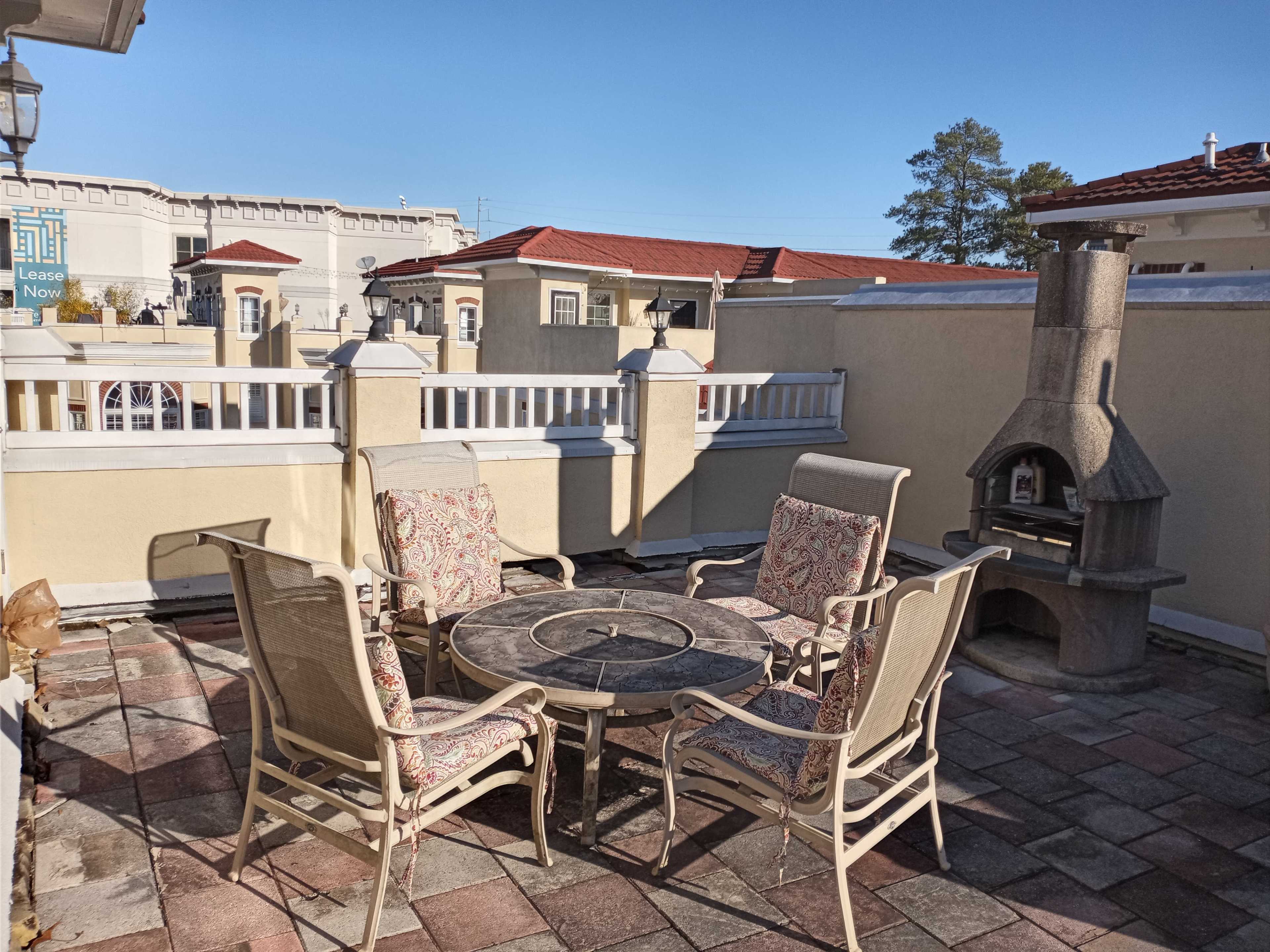 A patio area with four chairs and a round table is set around a stone fireplace, surrounded by a wall with a clear blue sky above.