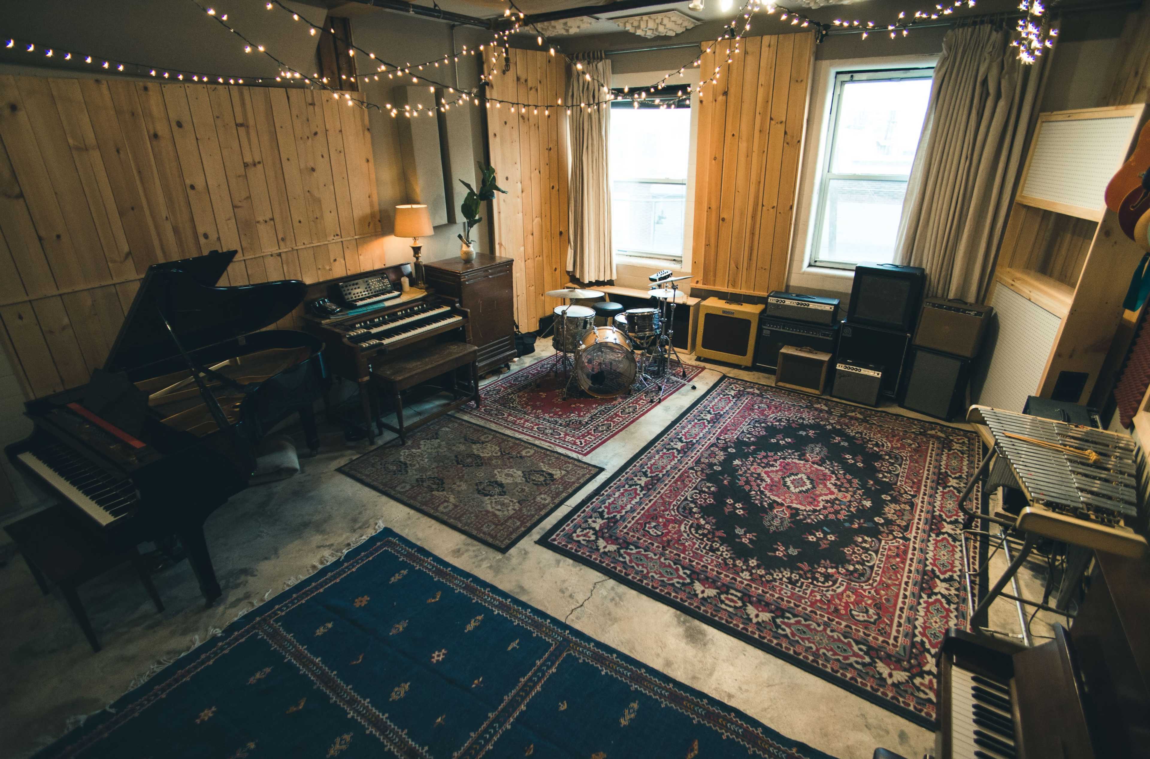 A music studio features various instruments, including a piano, drums, and amplifiers, arranged on patterned rugs with string lights overhead.