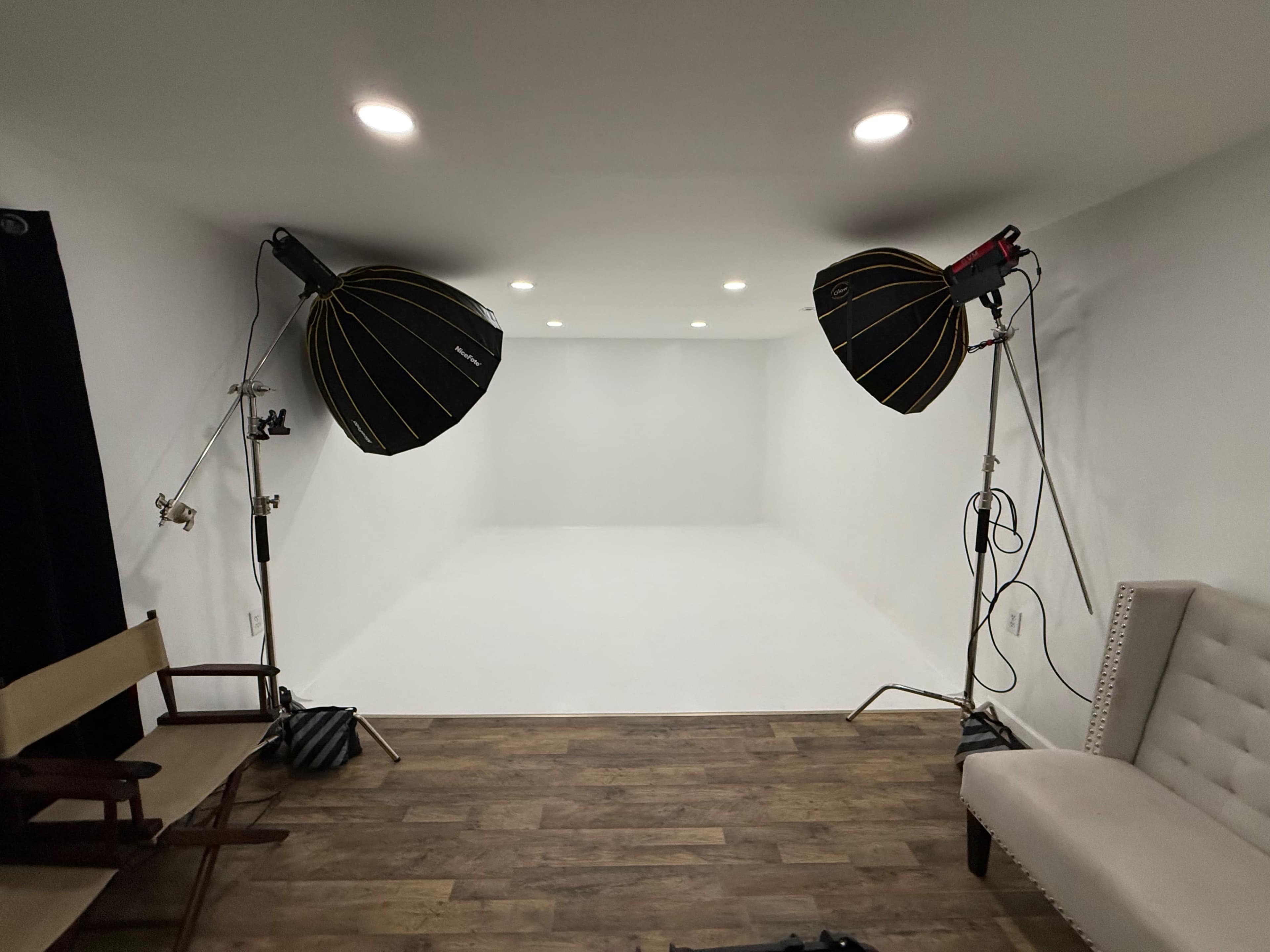 The image shows a photography studio with two large softbox lights positioned on either side and a white backdrop, along with a couple of chairs on a wooden floor.