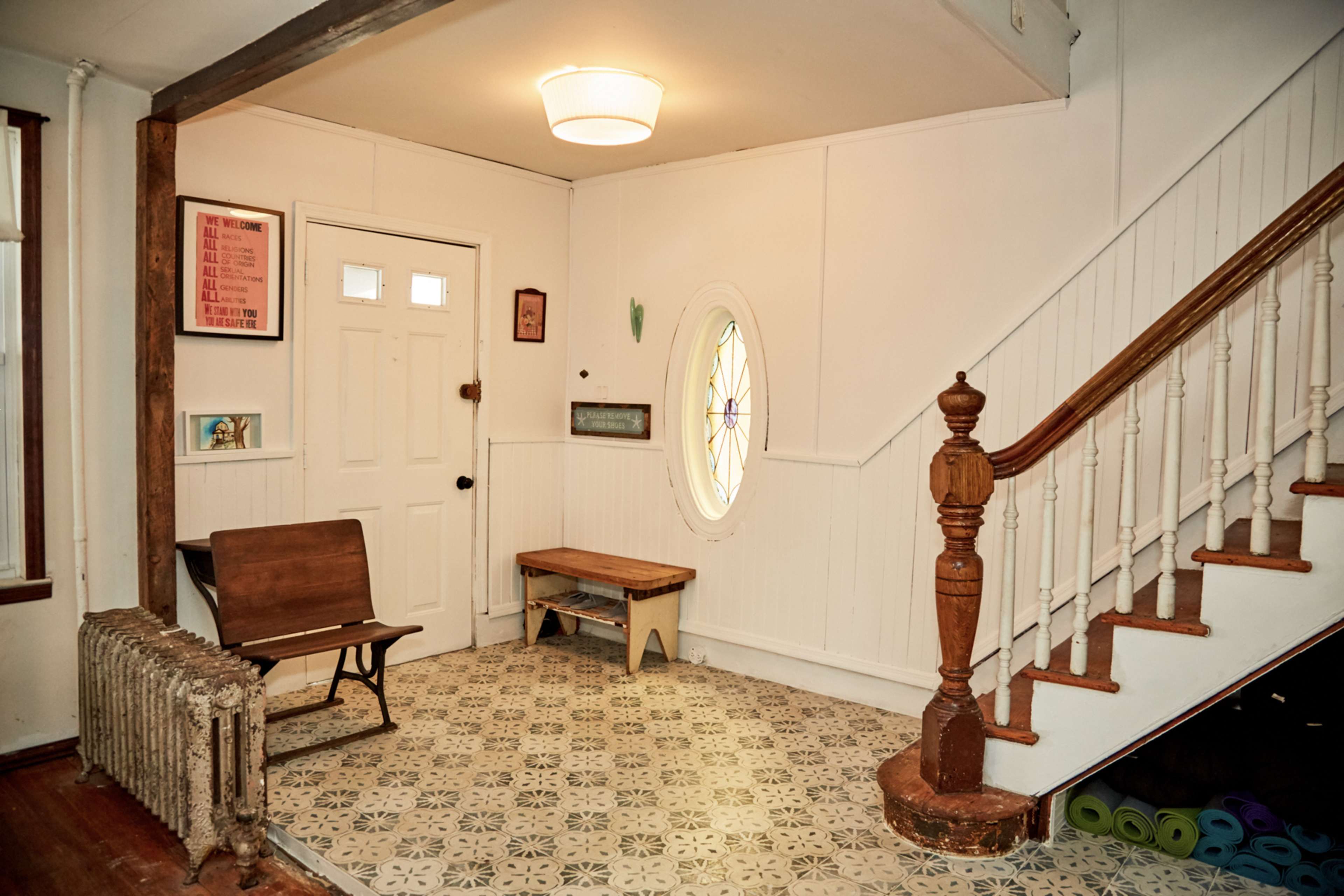 A hallway with a staircase, an oval window, a wooden bench, and a decorative radiator on the floor.