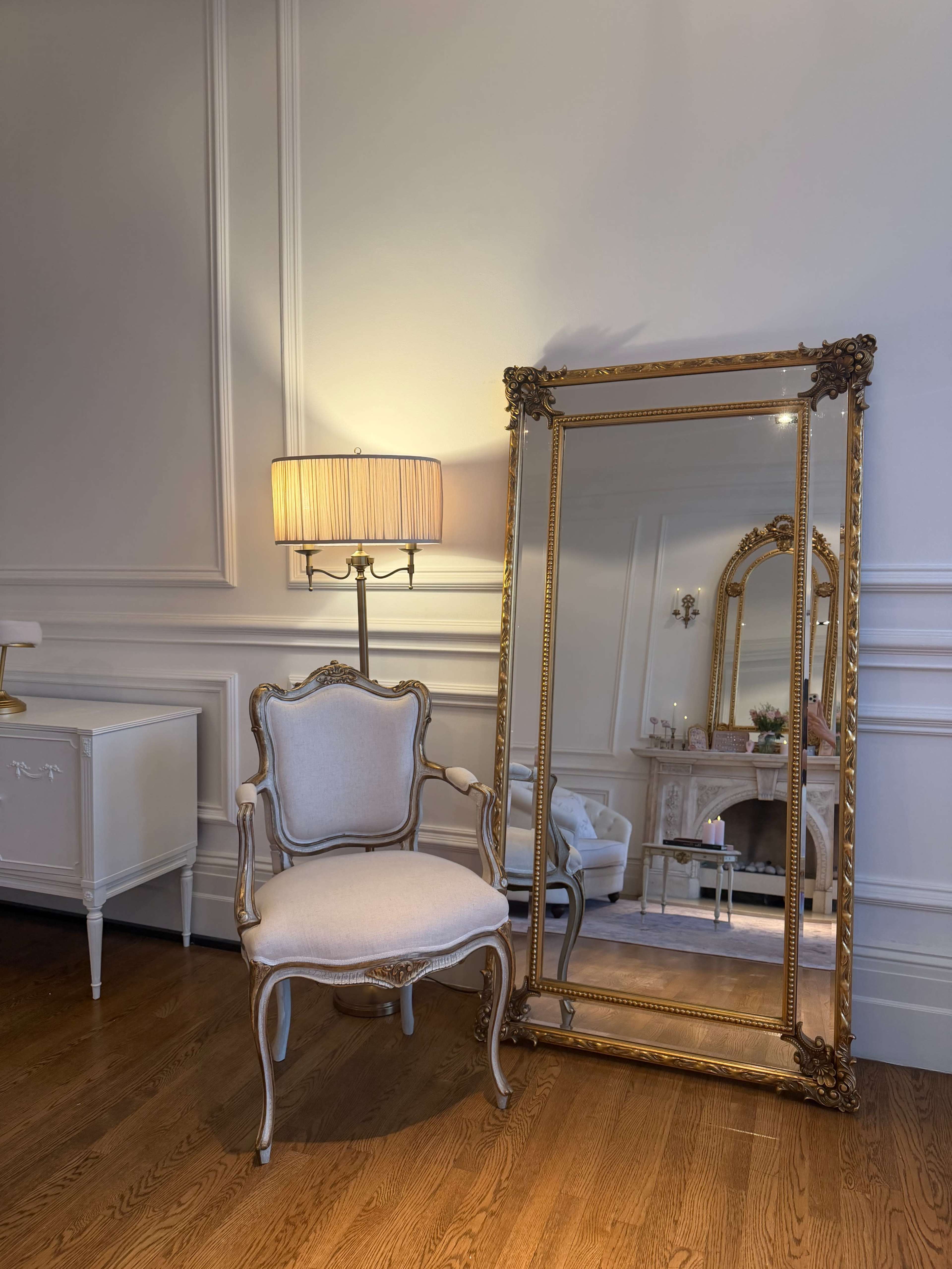 The image shows an elegant room featuring a vintage chair next to a large, ornate mirror and a lamp with a pleated shade.