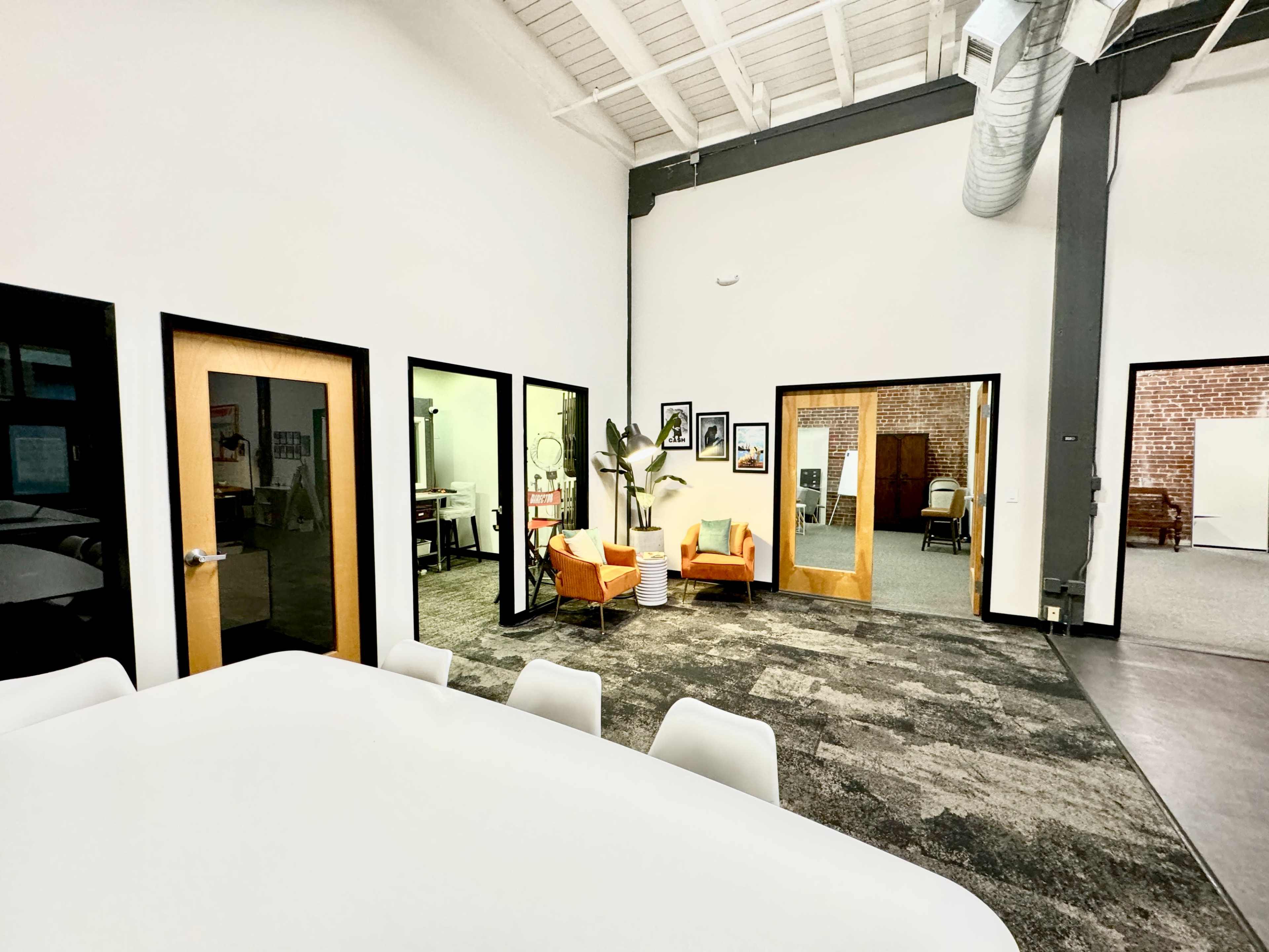 The image shows a spacious office interior featuring a large white table, several doorways leading to adjacent rooms, a seating area with orange chairs, and a backdrop of exposed brick walls.