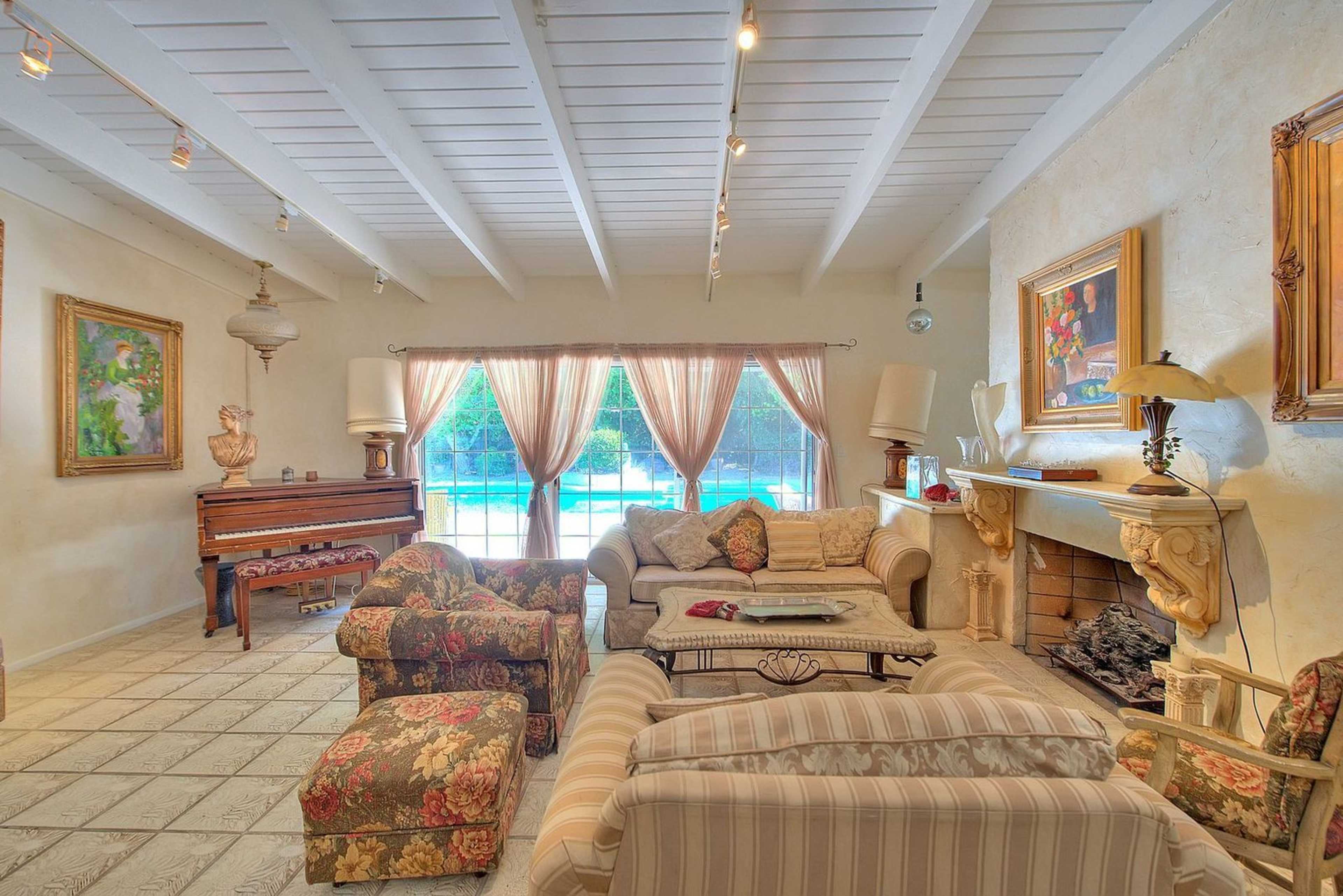 A cozy living room featuring floral-patterned sofas, a piano, and large windows overlooking a pool area.