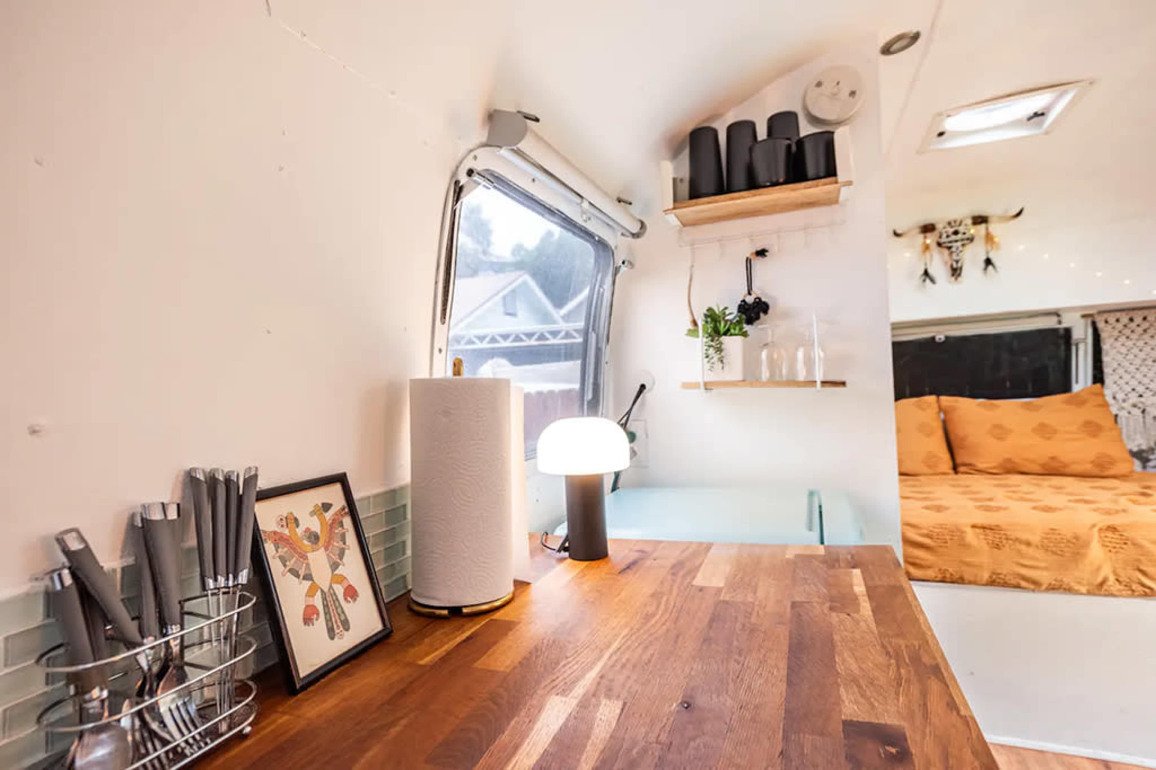 The image shows the interior of a compact living space with a wooden table, a lamp, and shelves displaying decorative items and plants.