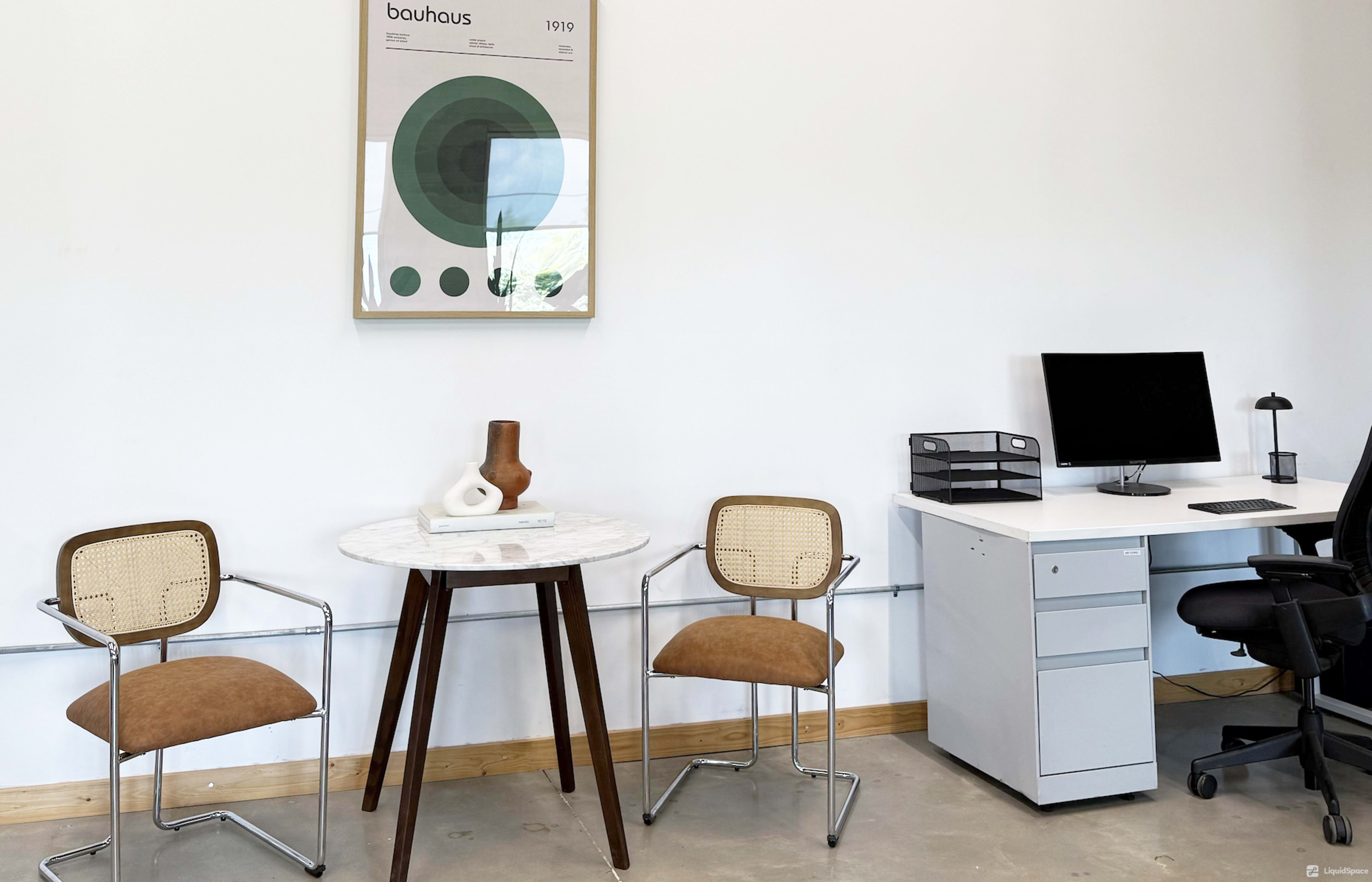 The image shows a minimalist office space featuring two chairs, a small round table with a decorative piece, and a desk with a computer monitor and office supplies.
