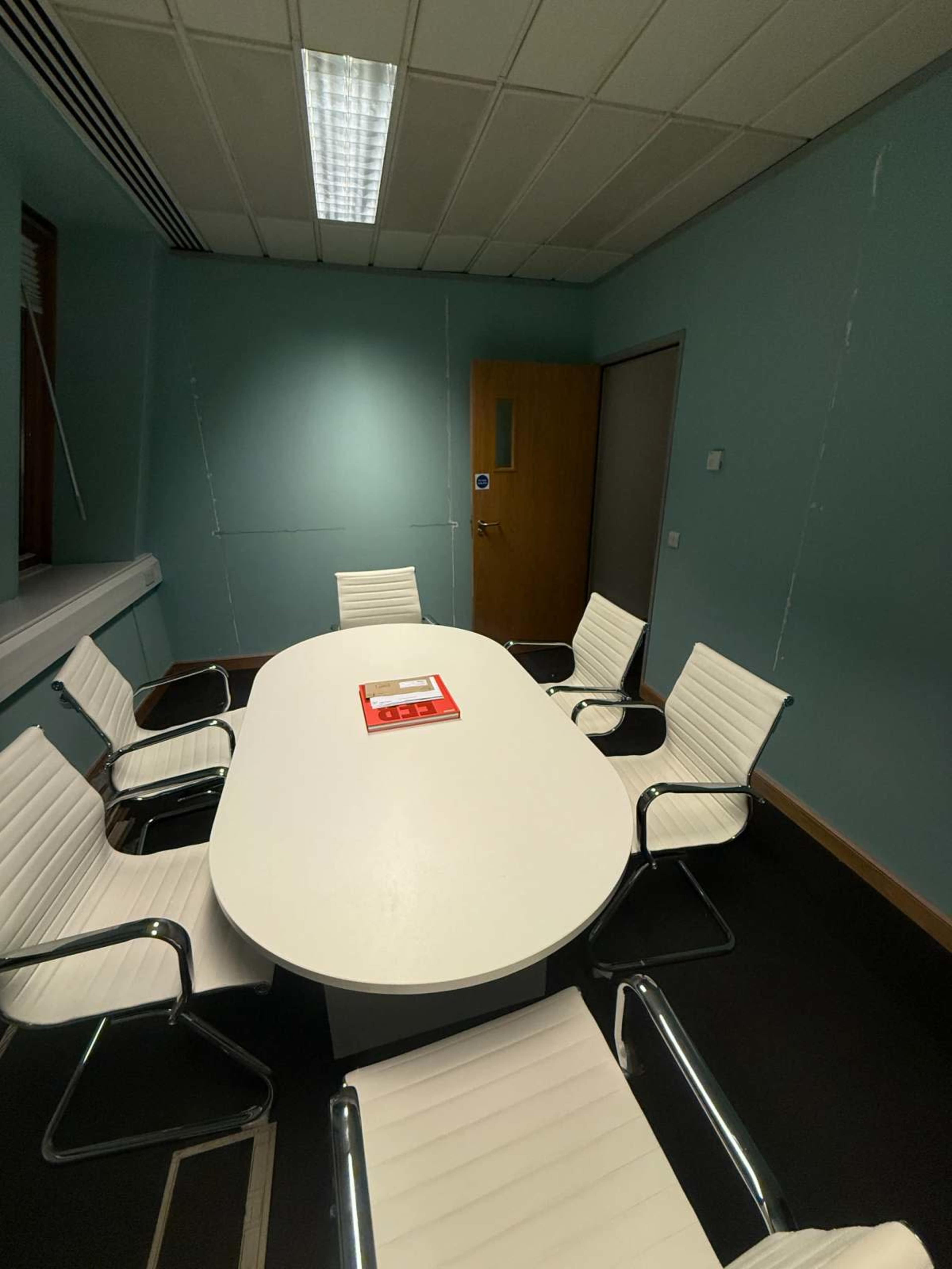 The image shows a conference room with a large oval white table surrounded by six white chairs, positioned against a light blue wall.