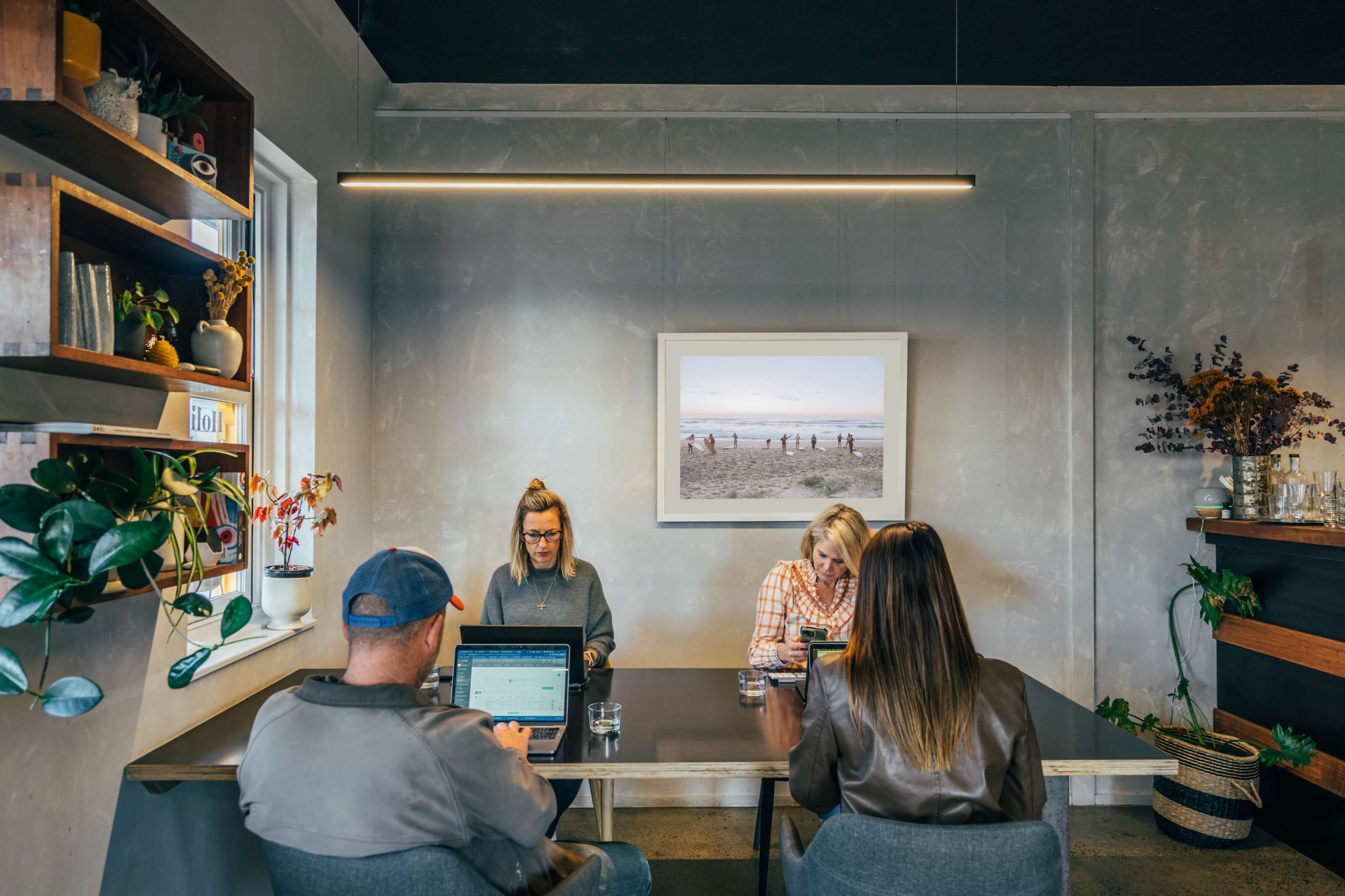 Four people are seated around a table in a modern room with a large window displaying a beach scene, while plants and art adorn the walls.