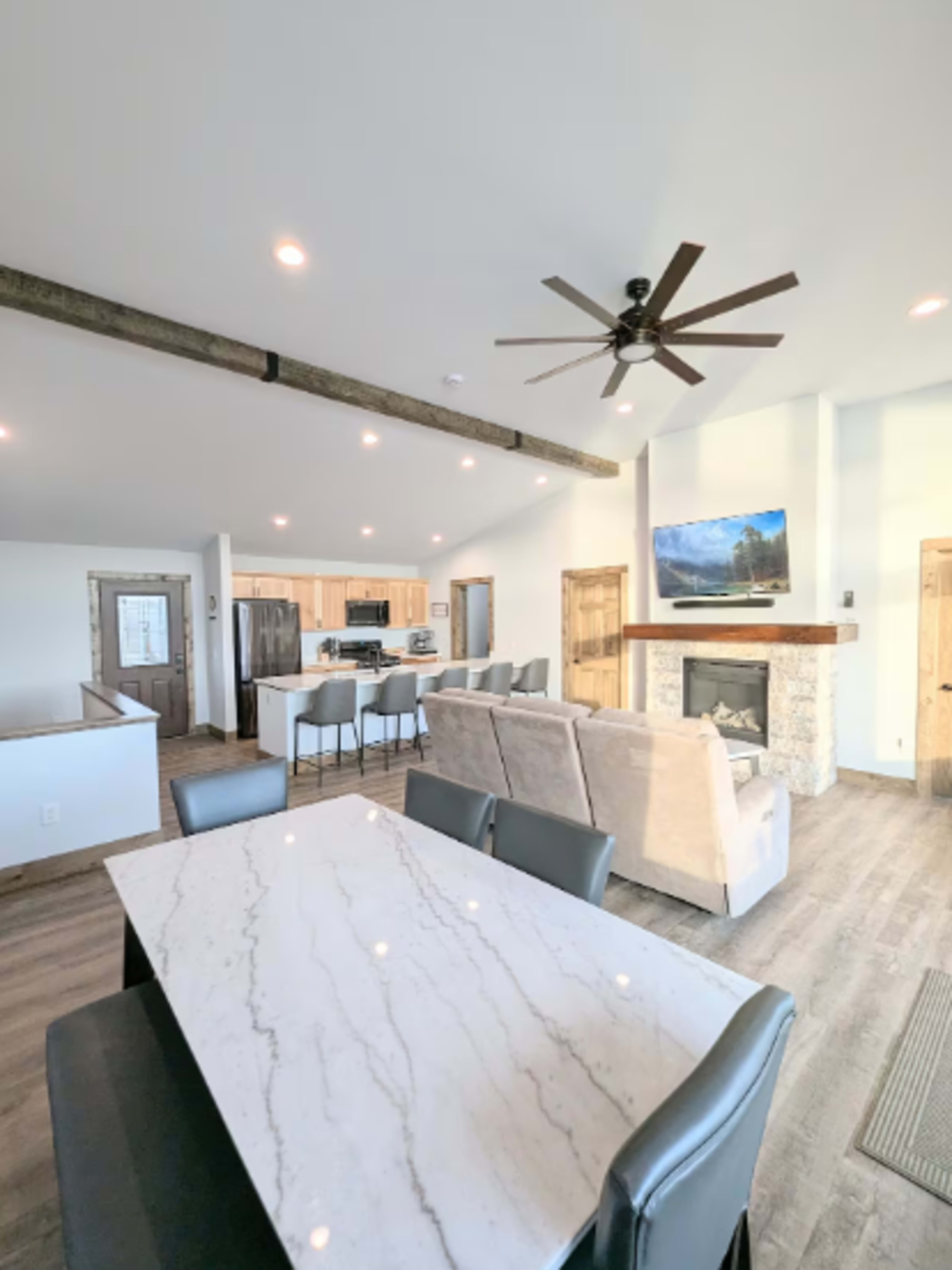 The image shows a modern open-concept living area with a kitchen, dining space, and a living room featuring a fireplace and a mounted television.