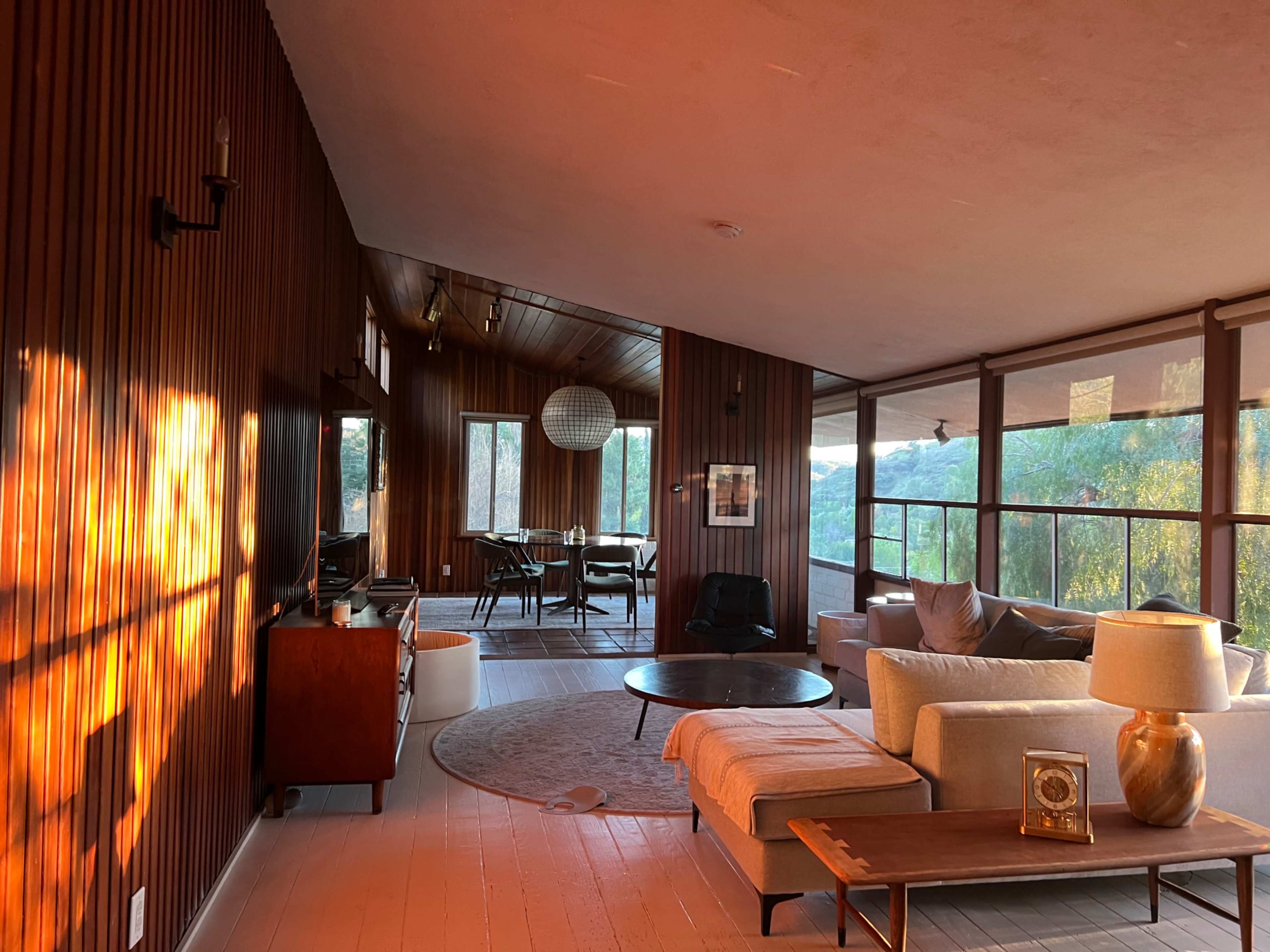 The image shows a modern interior living space with wooden walls, large windows, and a mix of seating arrangements.