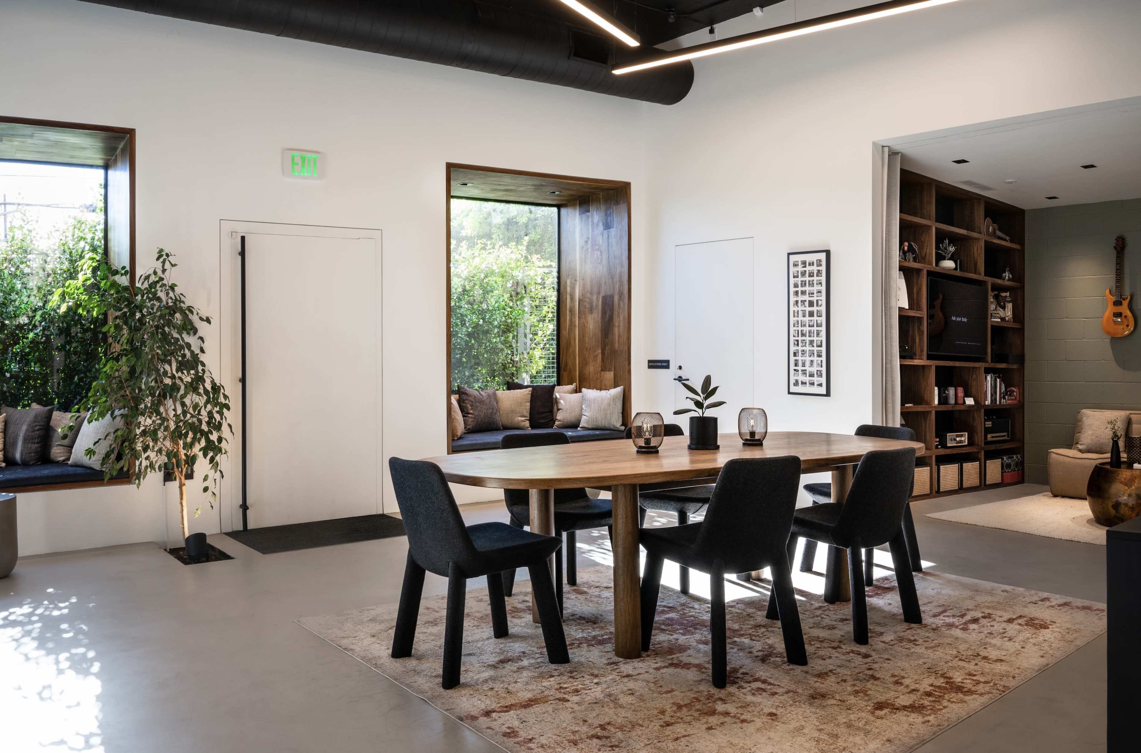 The image shows a modern dining room with a wooden table, black chairs, and large windows that provide natural light and views of greenery.