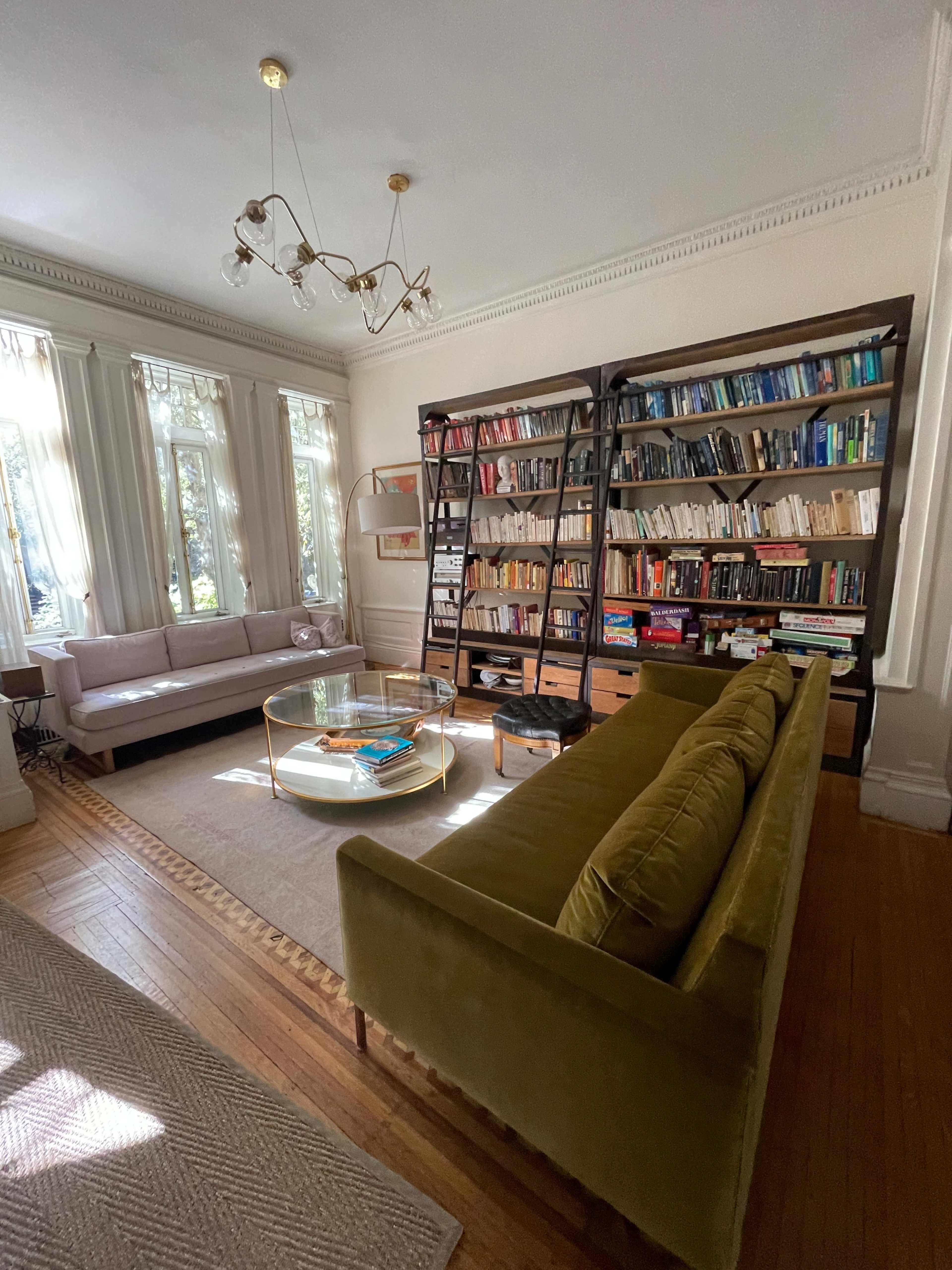 A cozy living room features a green sofa, a glass coffee table, a wall of bookshelves filled with books, and large windows allowing natural light to enter.