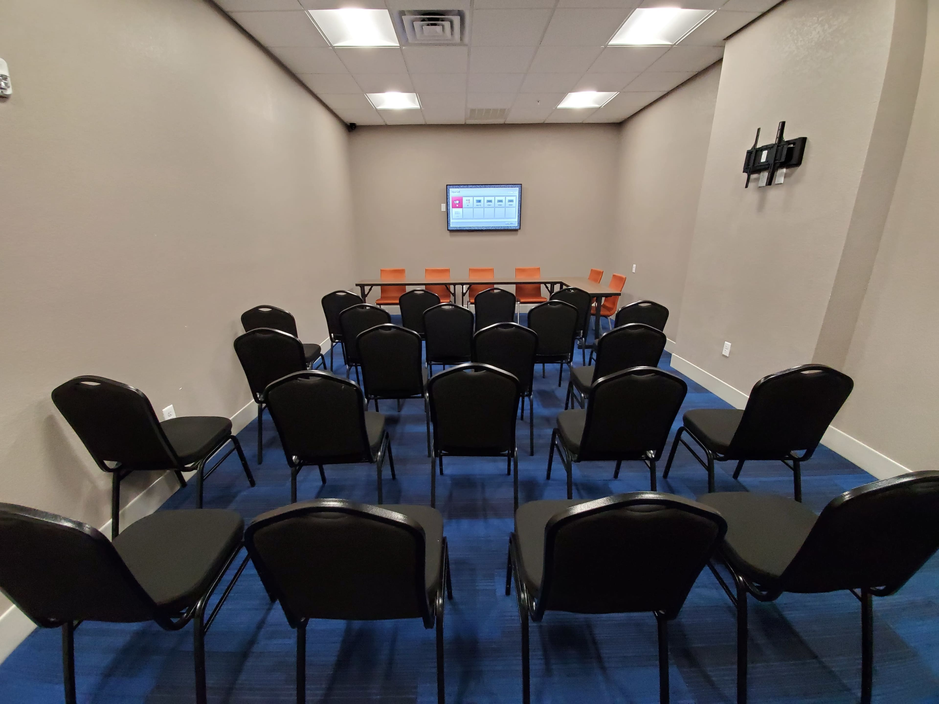 A meeting room is arranged with rows of black chairs facing a wall-mounted screen and a long table with orange chairs.