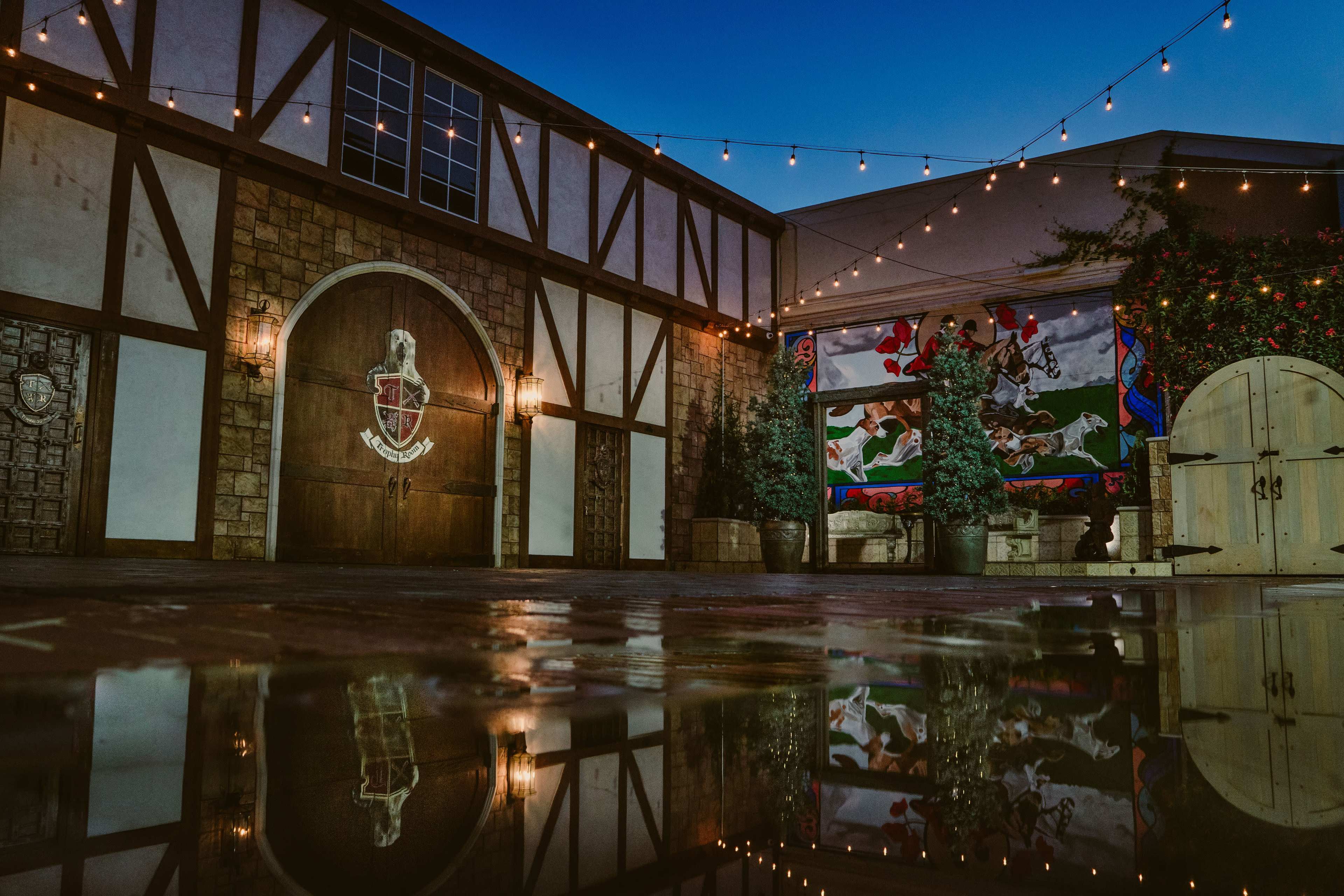 The scene features a rustic courtyard with a stone wall, a large wooden door, and a mural, all illuminated by string lights at dusk, reflecting in a puddle on the ground.