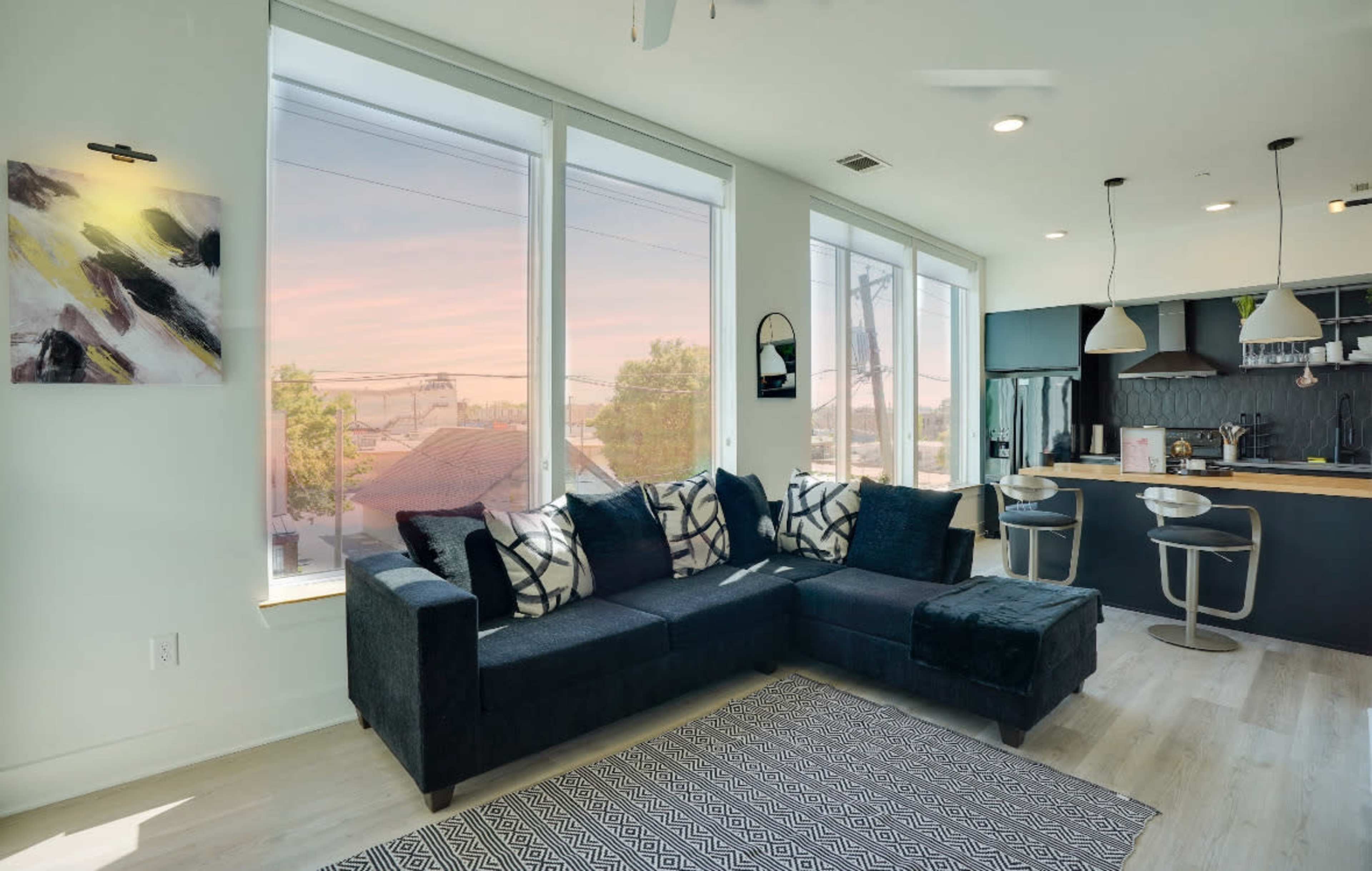 A modern living room features a dark sectional sofa with patterned throw pillows, large windows overlooking a sunset, and a kitchen area with bar seating.
