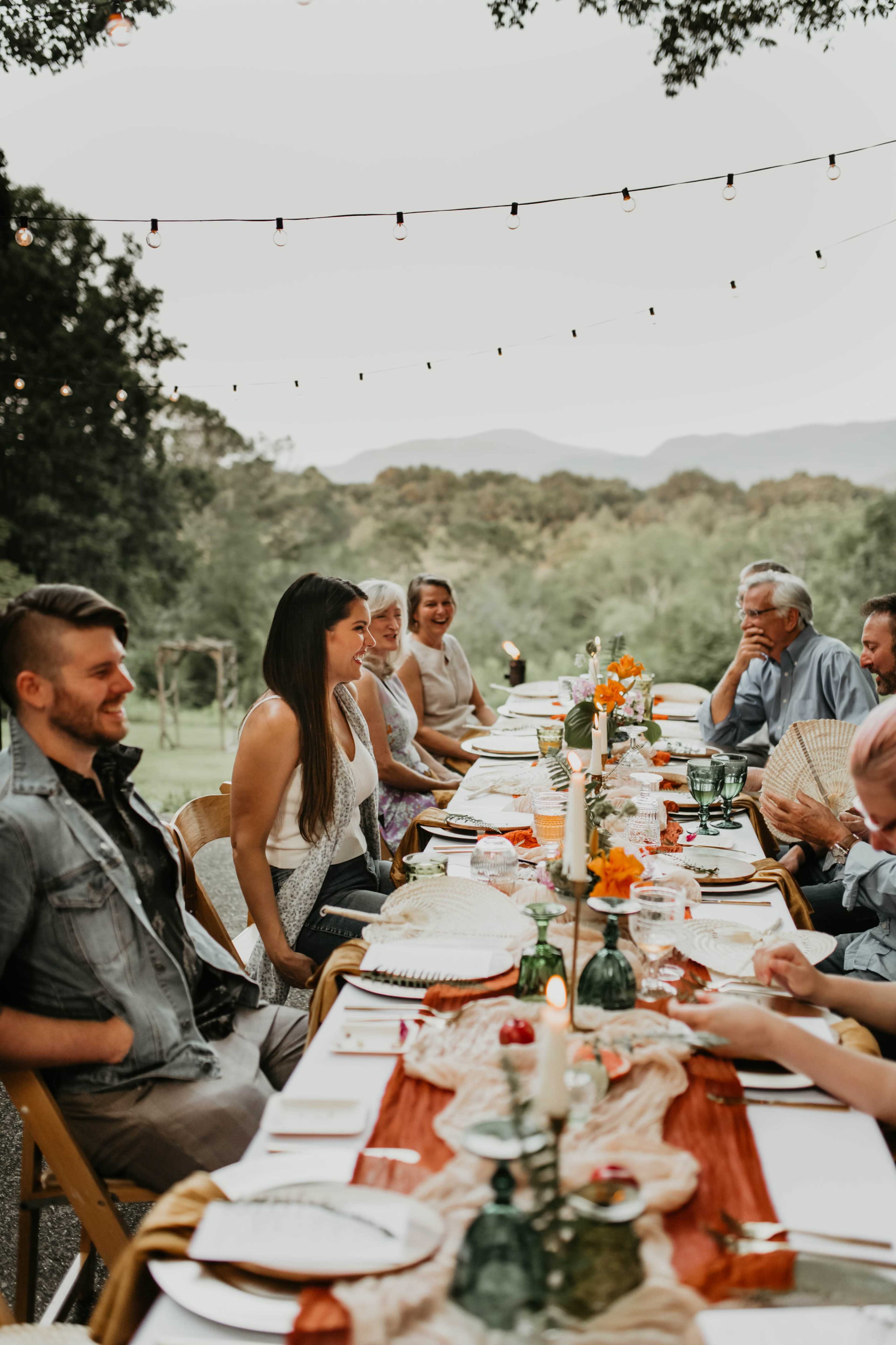 A long table is set for a dinner party outdoors, with multiple guests seated across from each other, surrounded by greenery and mountains in the background.