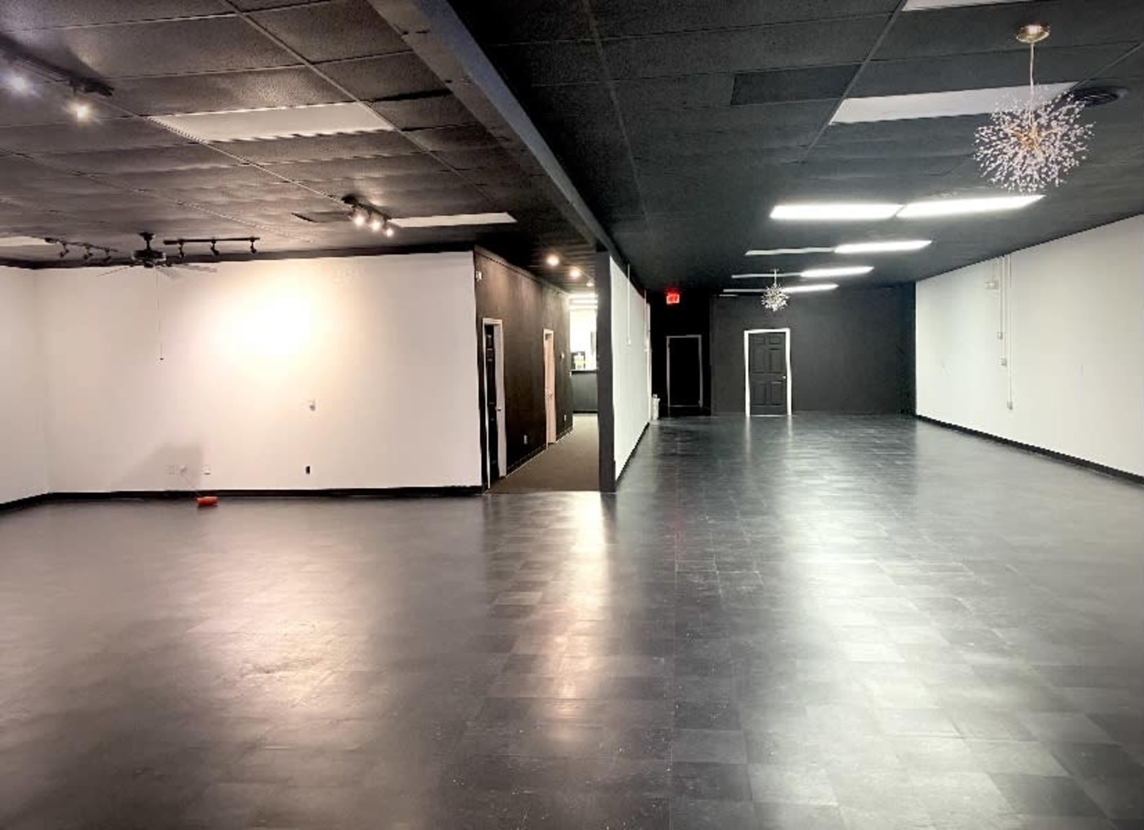 The image shows a spacious, empty interior room with black tiled flooring, white and black walls, and large overhead lights.