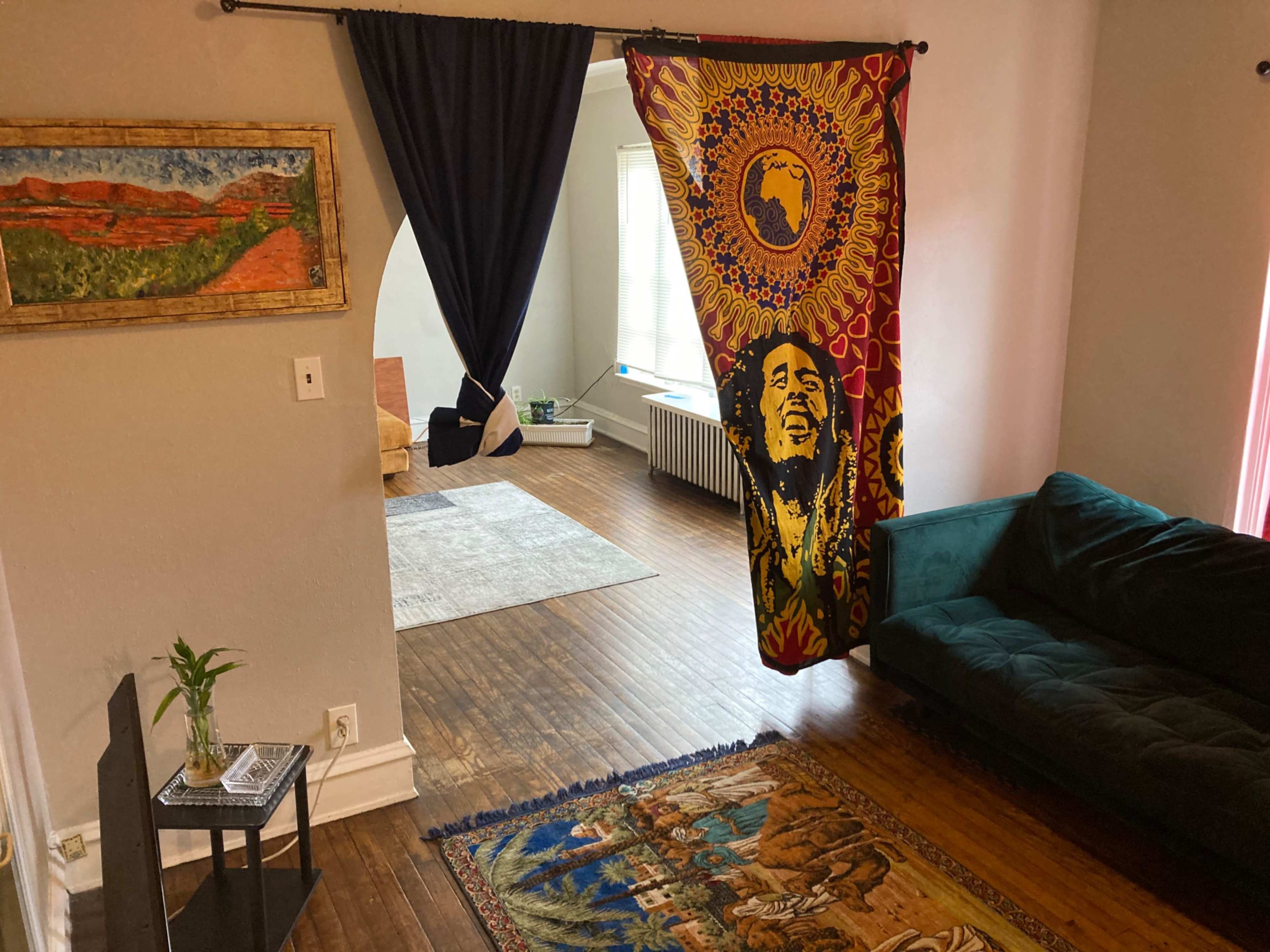 A living space divided by a vibrant tapestry featuring a figure, leading to another room with wooden floors and a sofa.