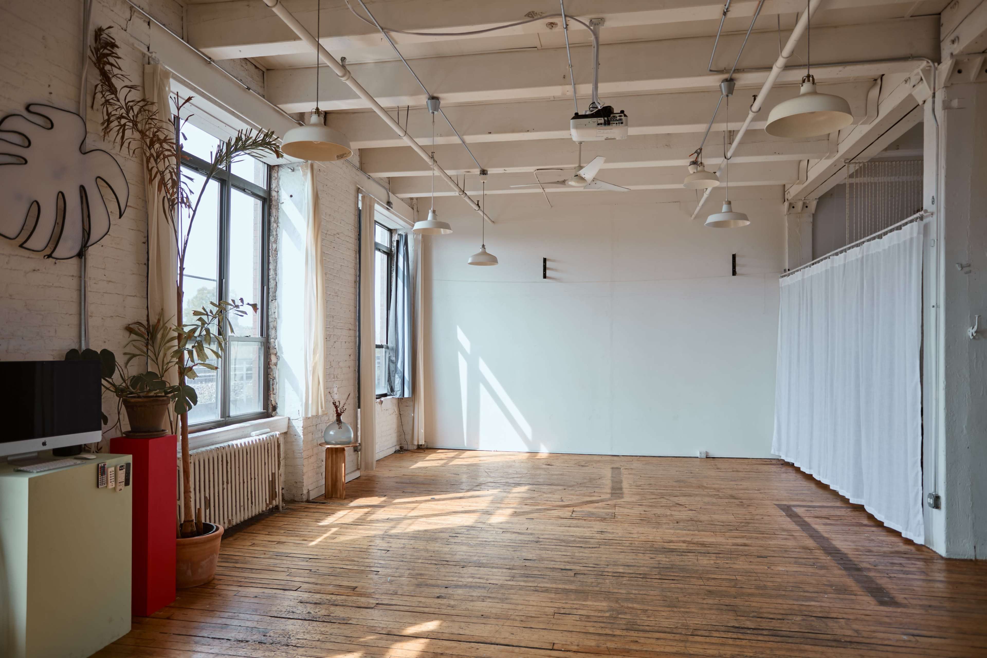 A spacious and well-lit interior features wooden floors, large windows, and a white wall adorned with plants and minimalist decor.