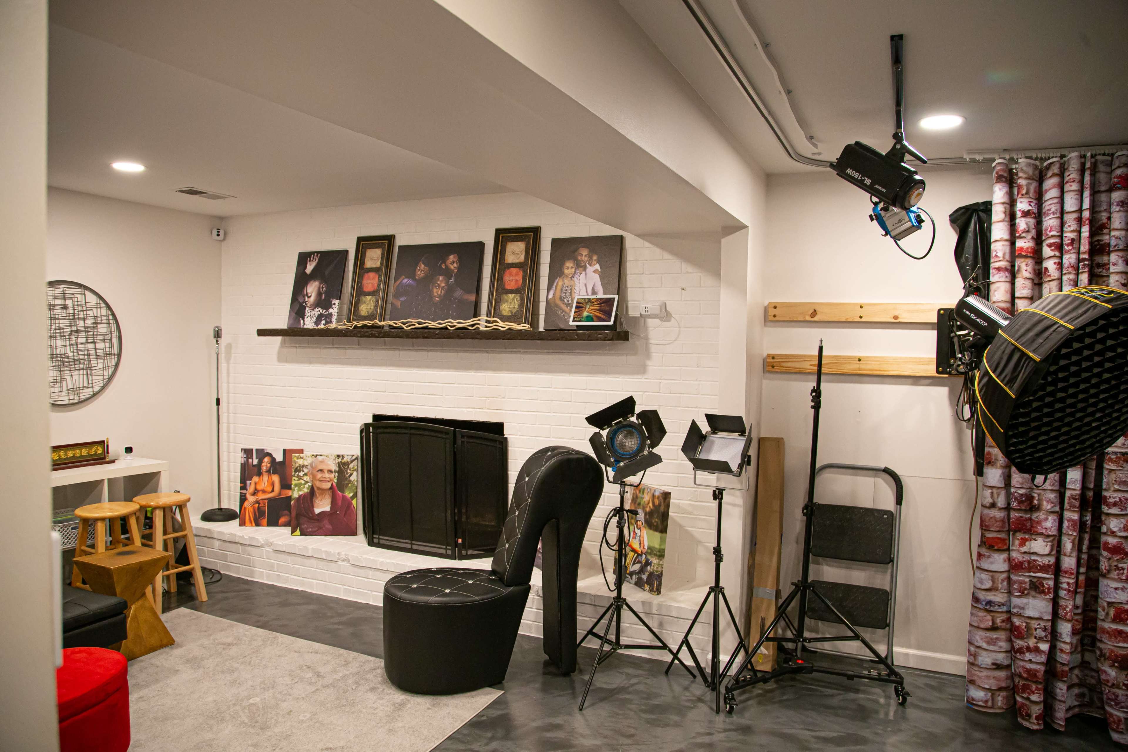 A studio setup featuring a white brick wall with a mantel, various photographs displayed above it, a large high-heeled shoe chair, and studio lighting equipment on the floor.