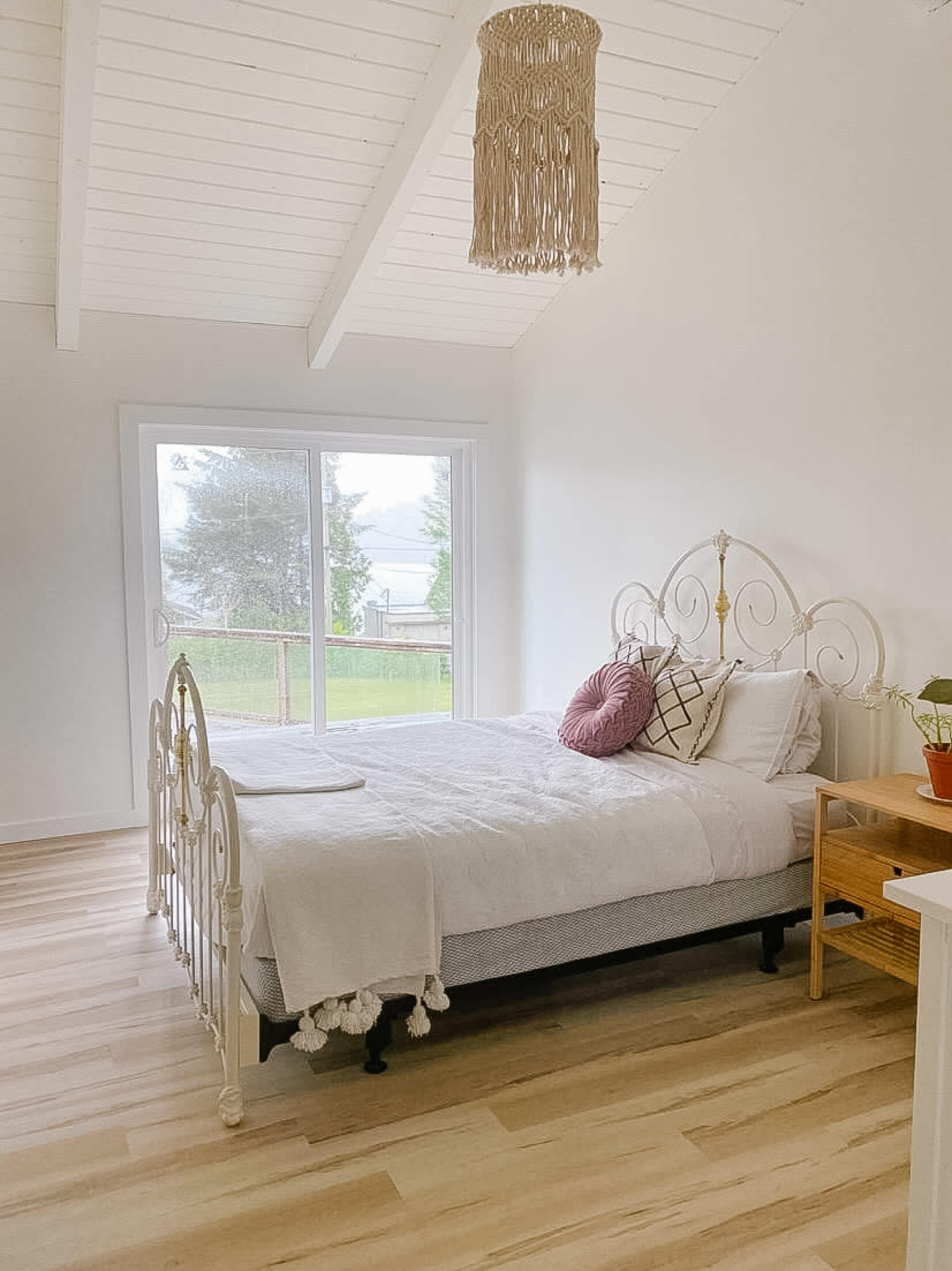 A bright bedroom features a bed with a white headboard, a pink pillow, and a large window overlooking a grassy area.