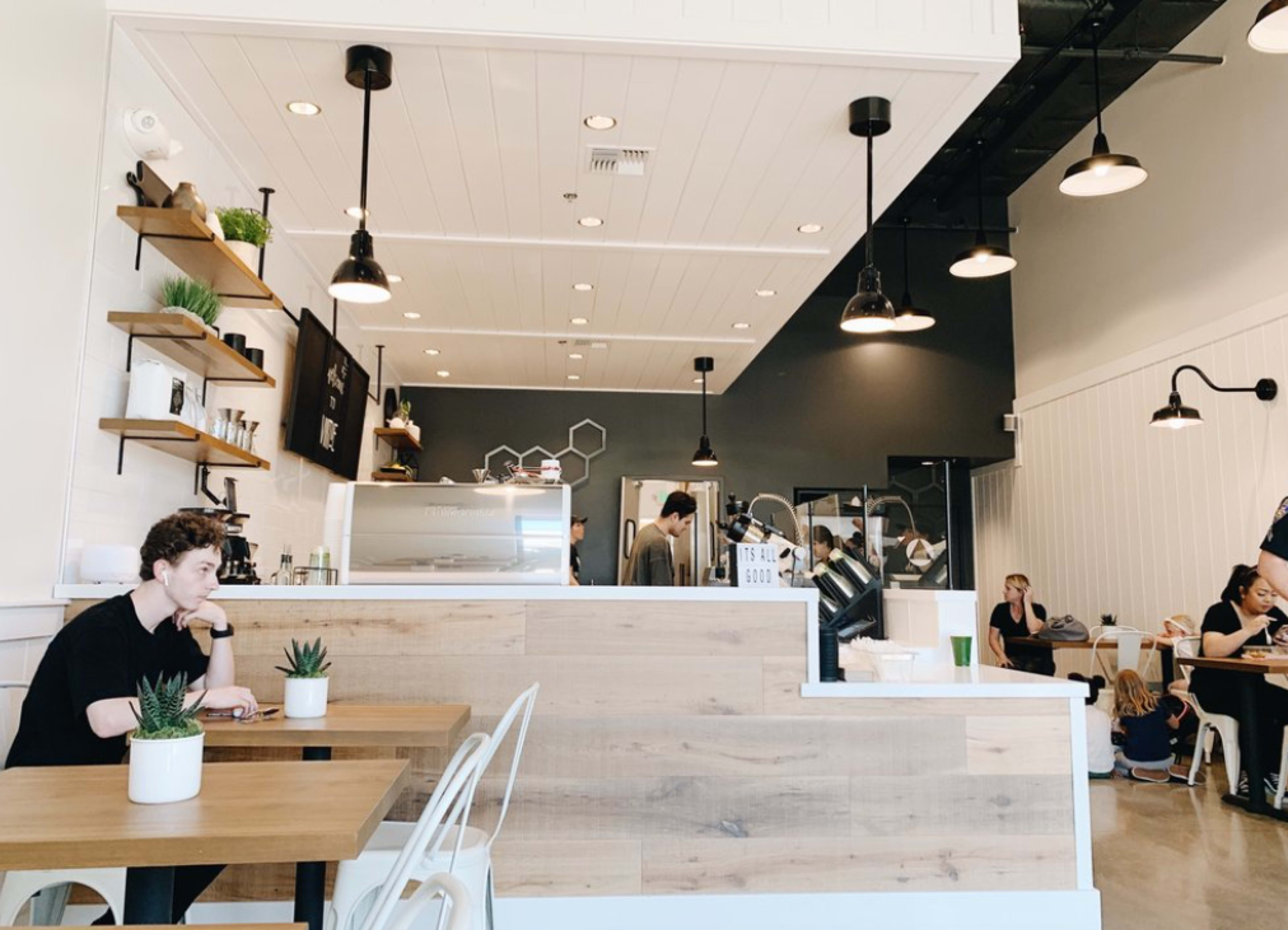 A coffee shop interior features a light wood counter, shelves with plants, and several customers seated at tables.