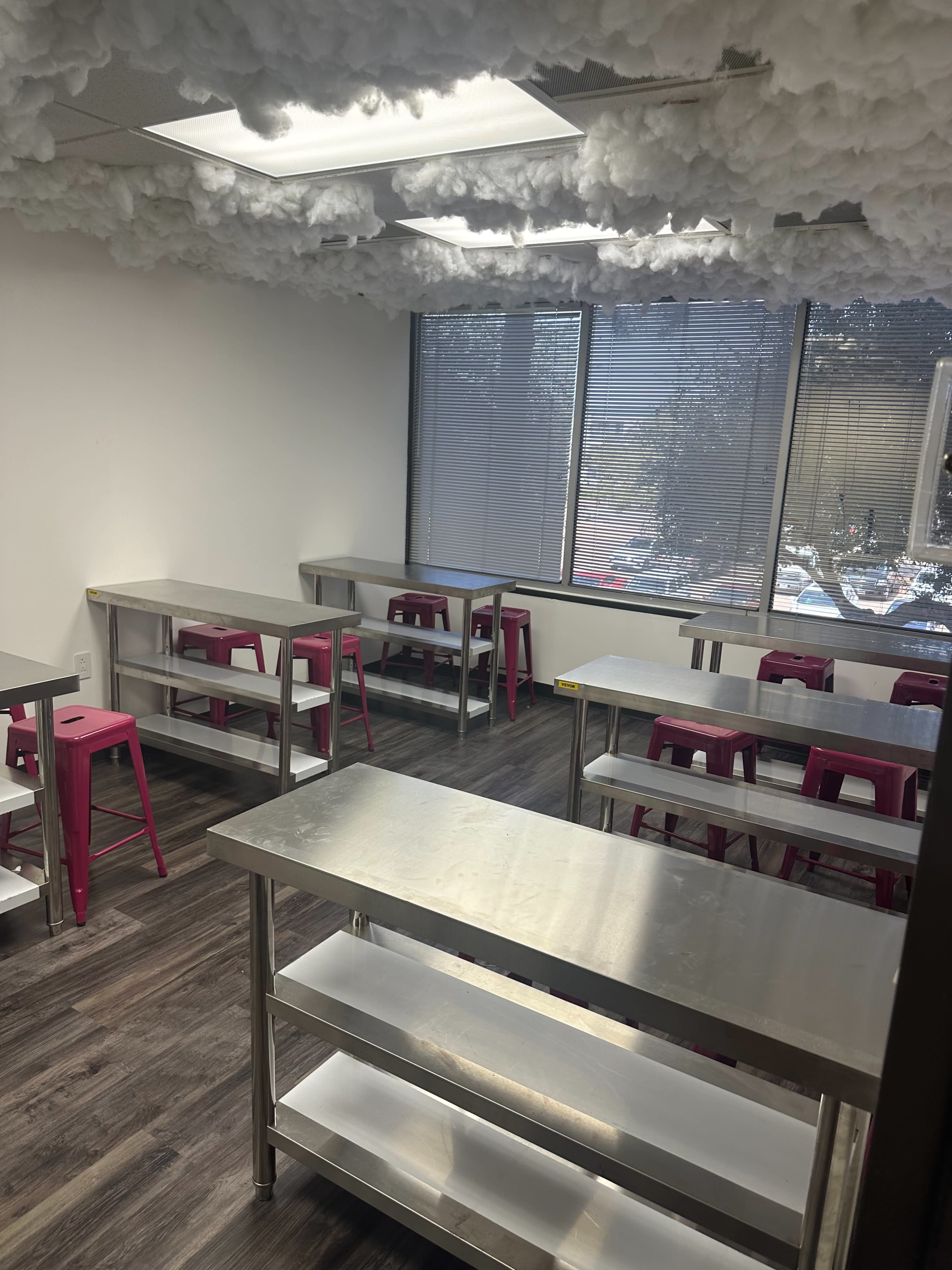 A classroom with stainless steel tables and pink stools, under a cloud-like ceiling, and large windows letting in natural light.