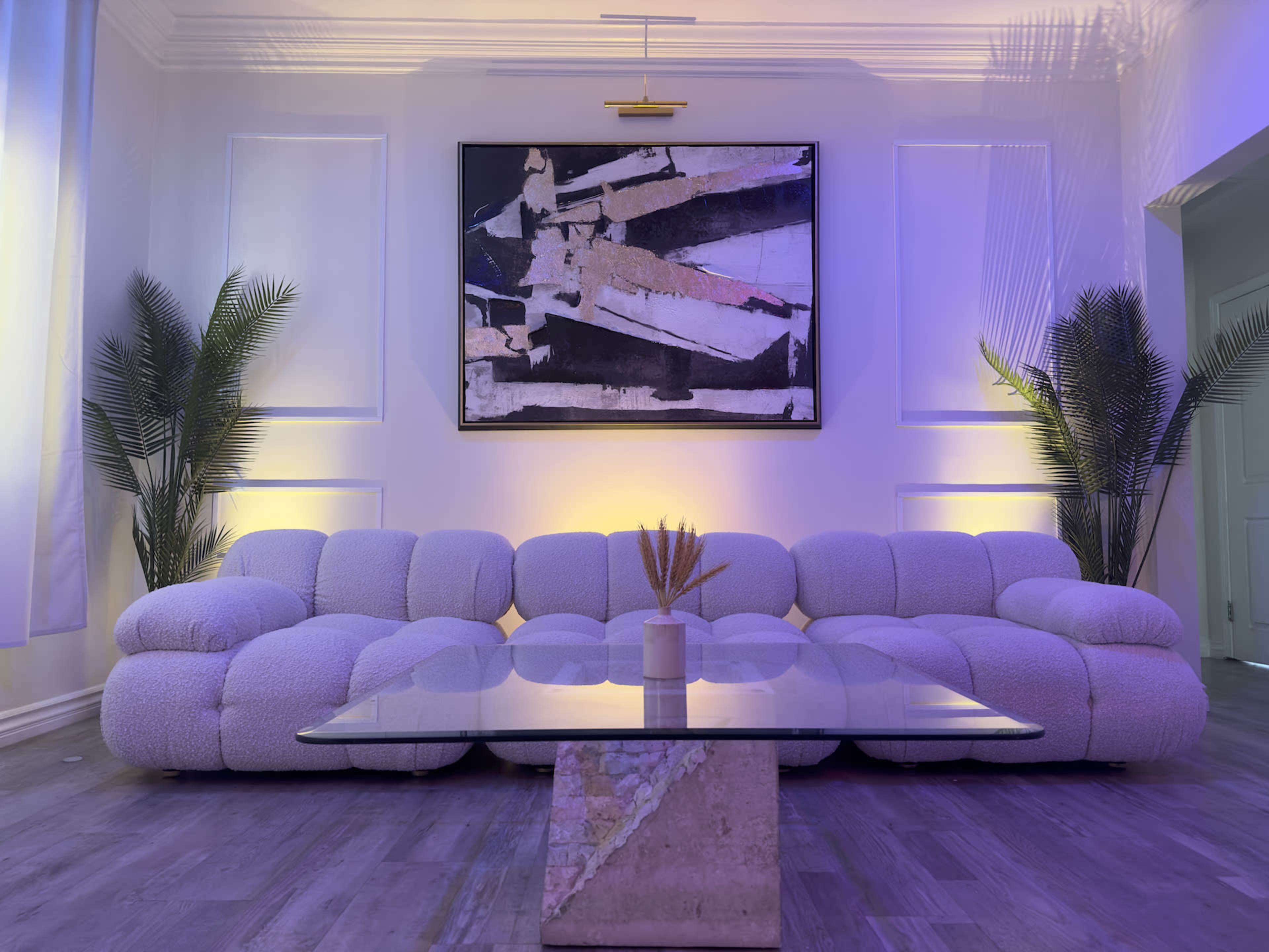 A modern living room features a plush, light-colored sofa, a glass coffee table, and an abstract painting on the wall, illuminated by soft lighting.