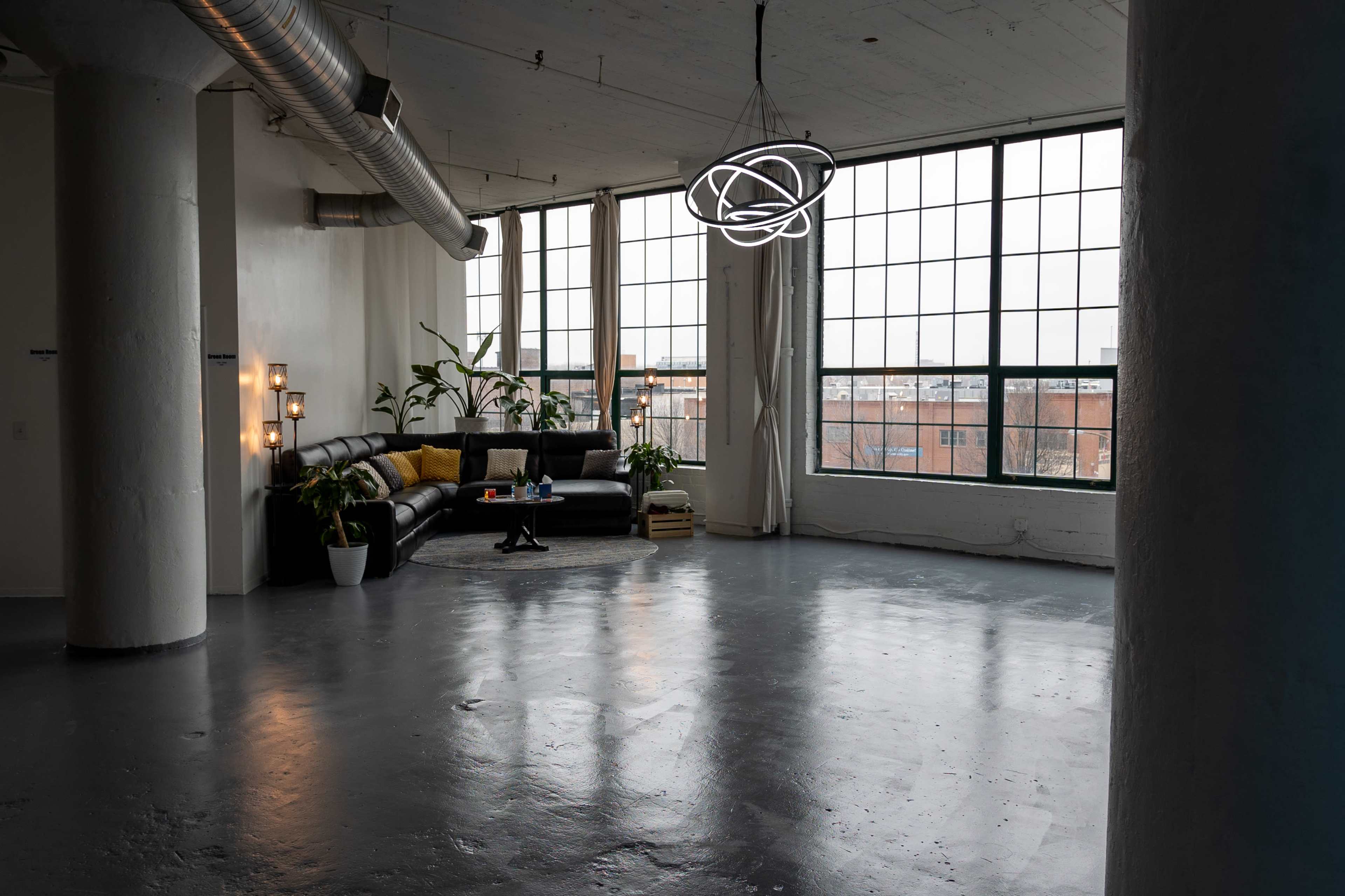 A spacious, minimalist living area with large windows, a black sofa, plants, and a modern light fixture.