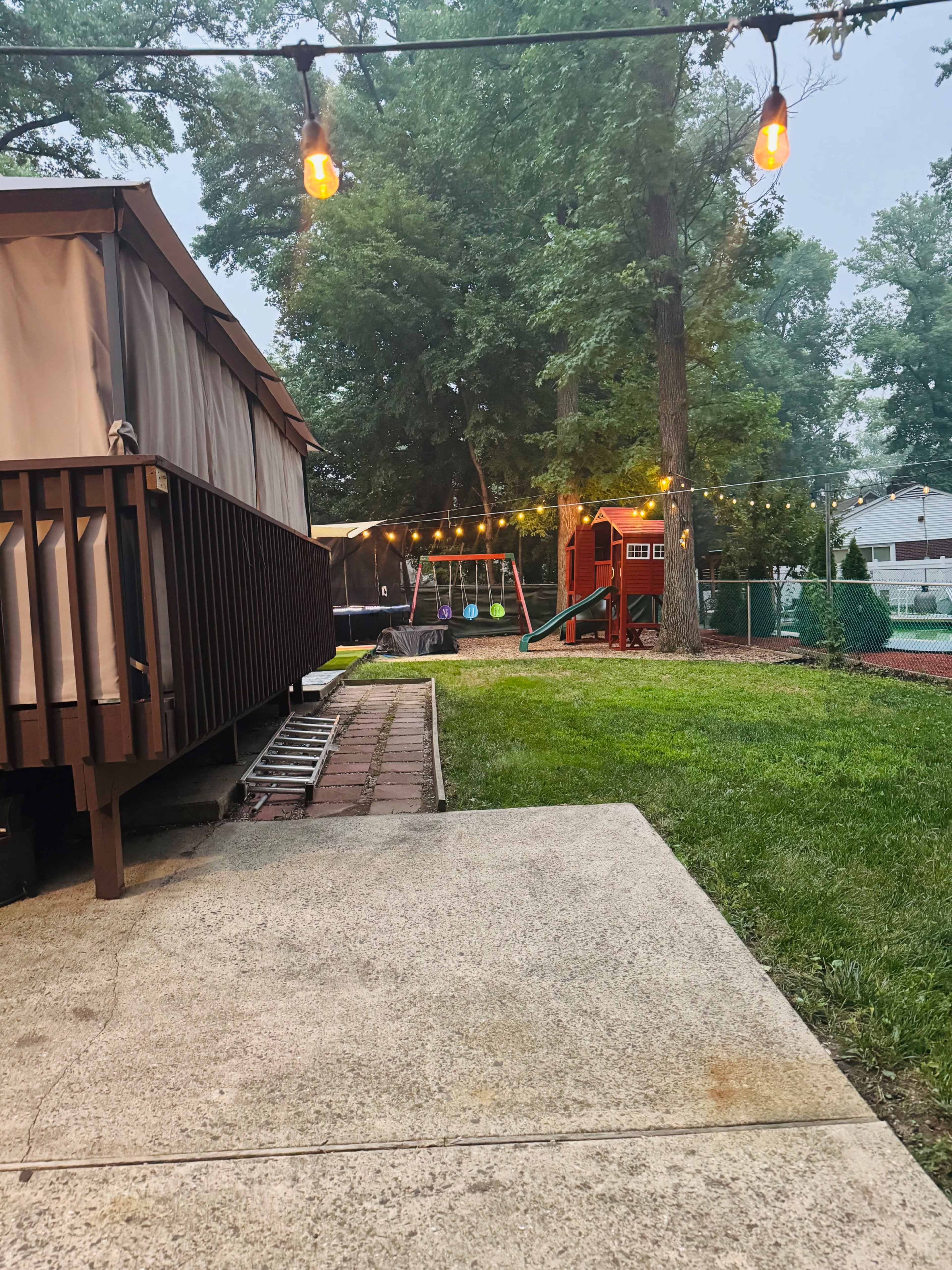 The image shows a backyard with a wooden deck, a playground set, and string lights hanging among the trees.