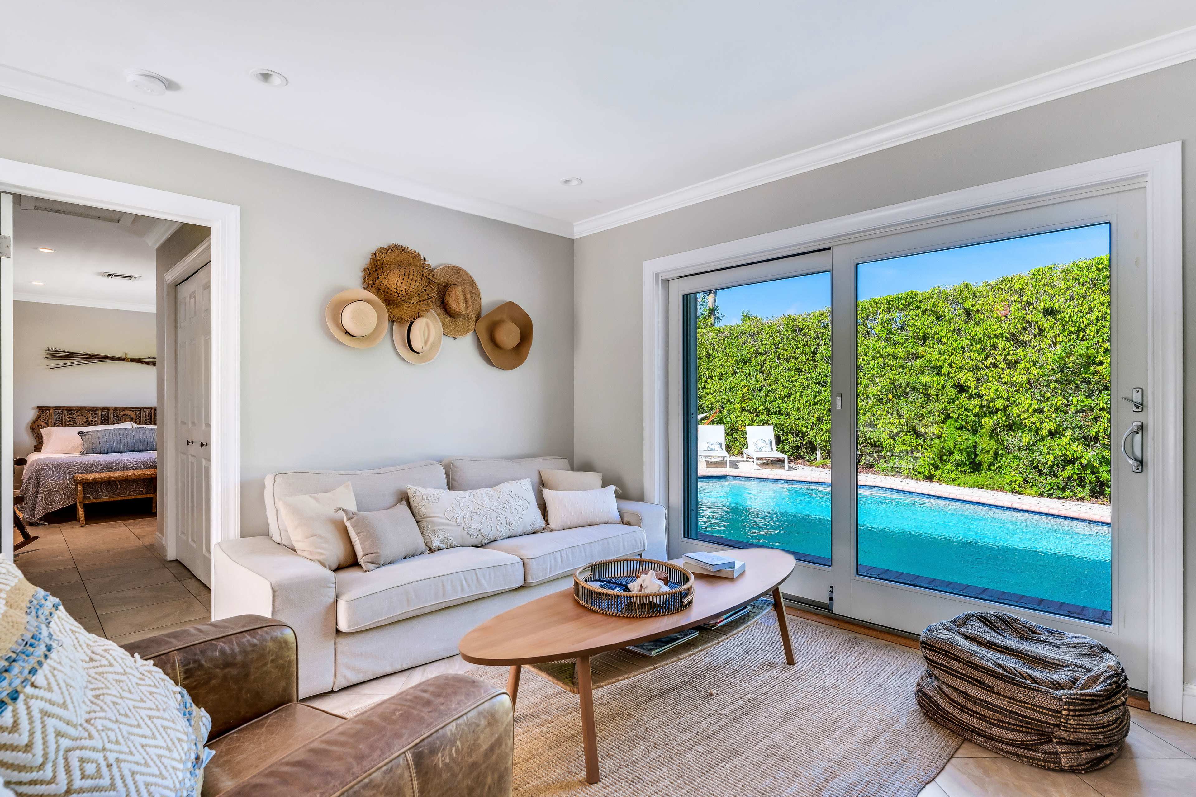 A modern living room with a beige sofa, coffee table, and large glass doors opening to a pool area surrounded by greenery.