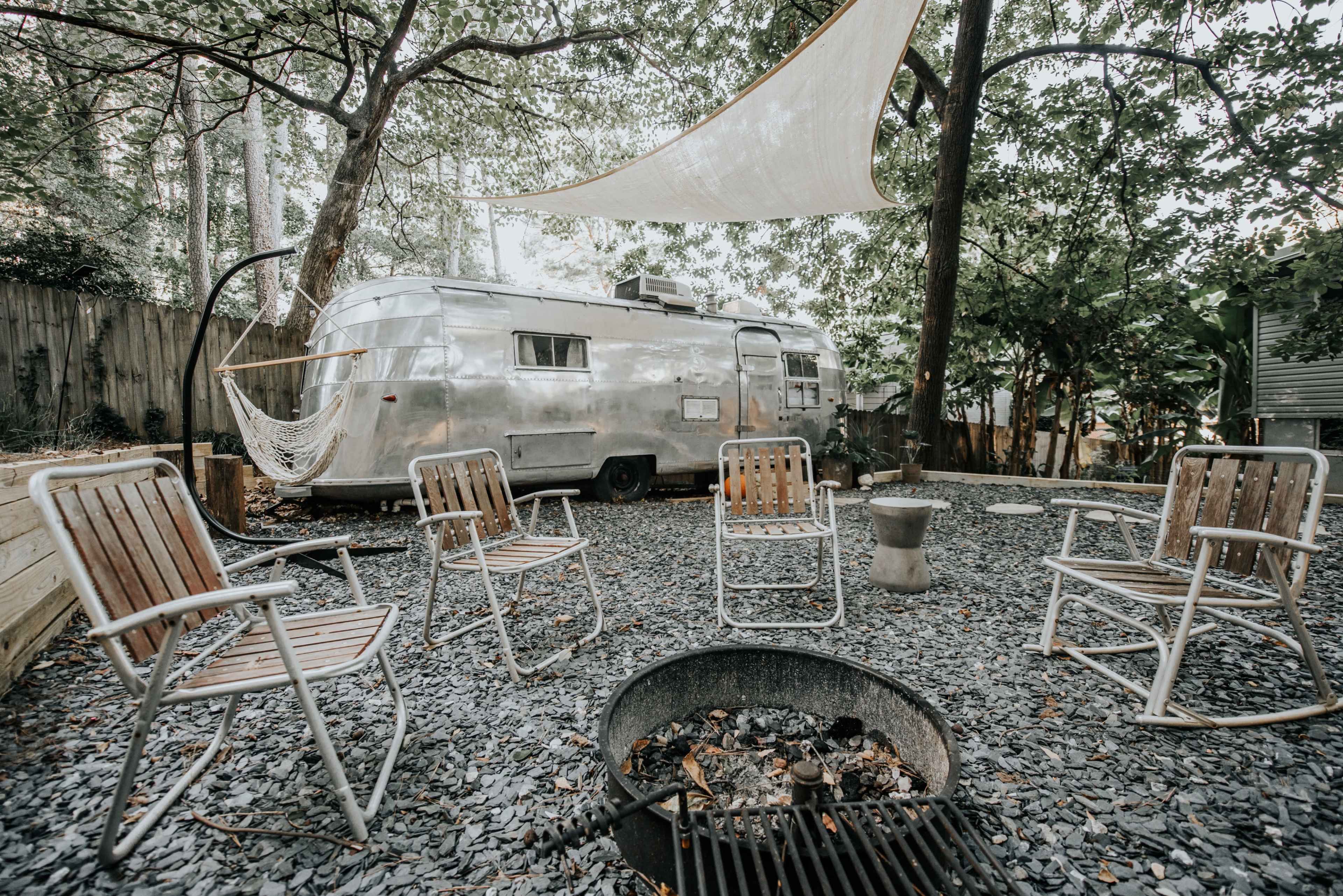 A circular fire pit surrounded by four wooden chairs is set in a gravel yard with a vintage Airstream trailer in the background.
