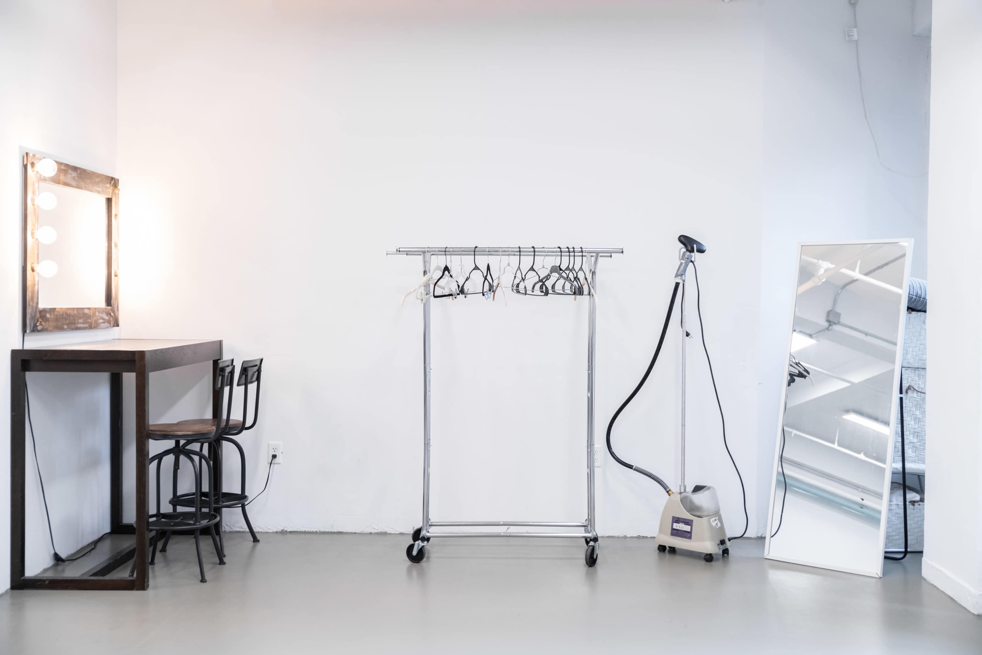 A minimalistic dressing room with a table and chairs, a clothing rack with hangers, a garment steamer, and a large mirror against a white wall.