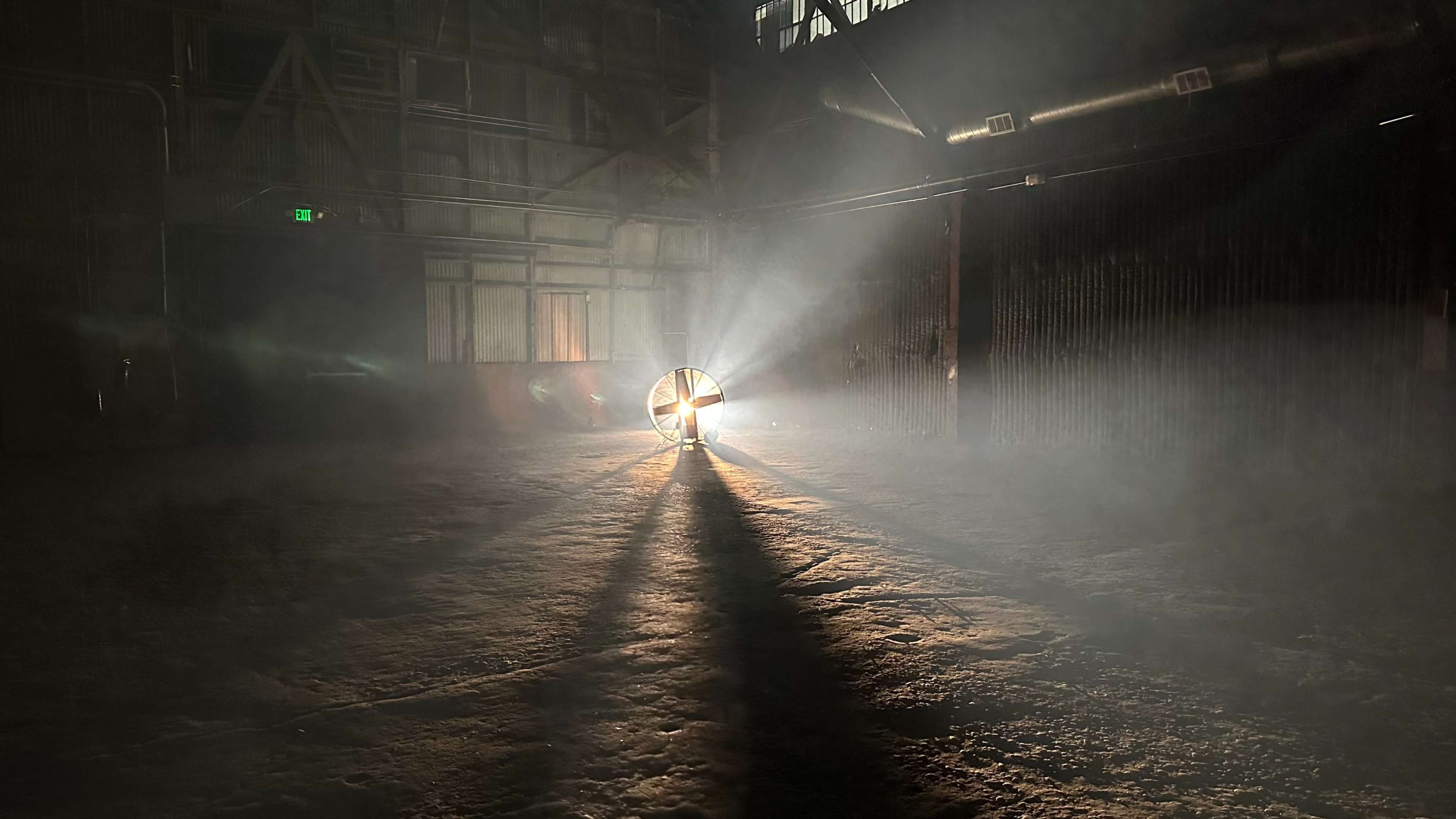 A spotlight beams through a foggy, dimly lit industrial space, casting long shadows on the ground.