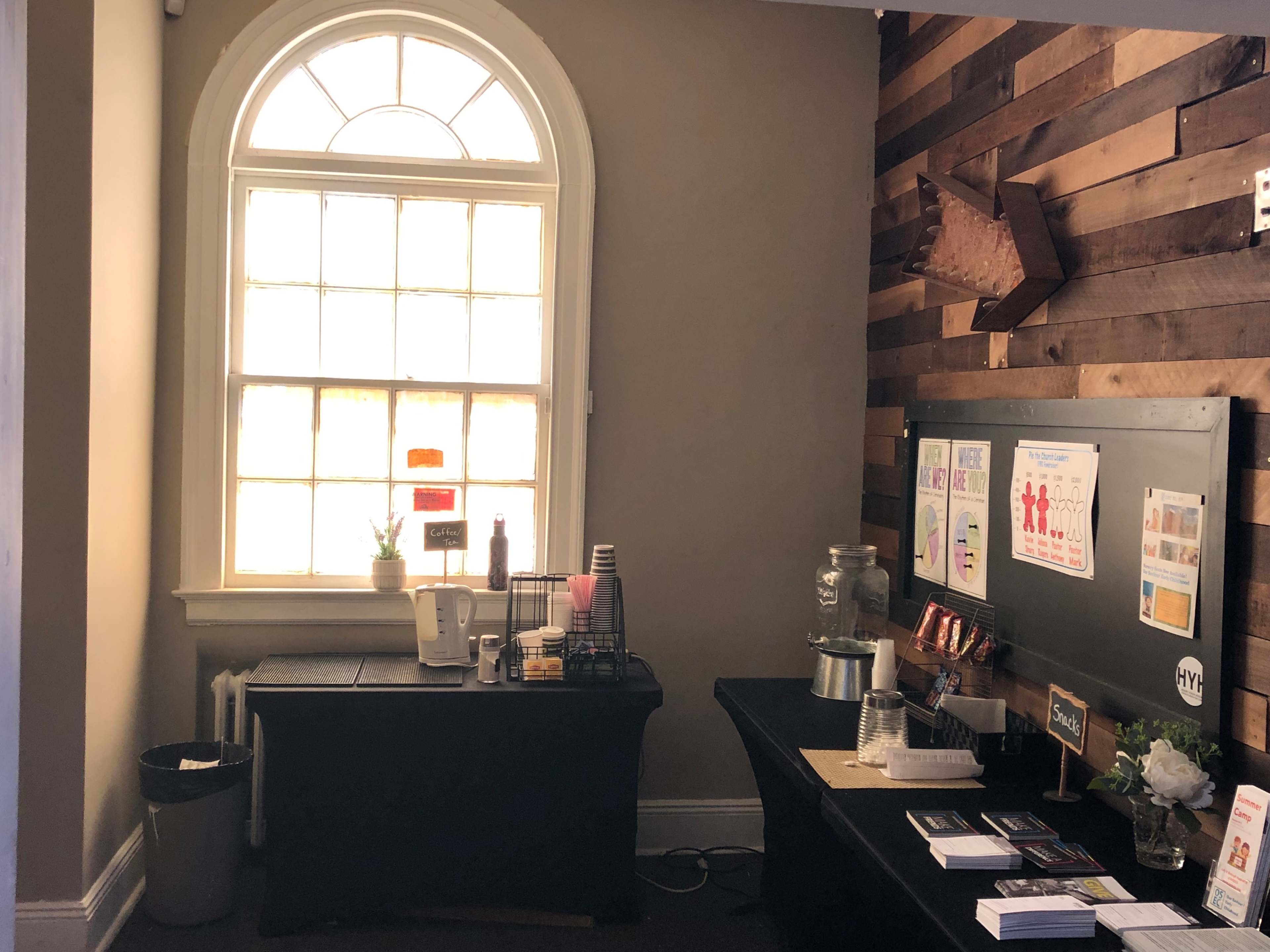 A reception area featuring a large window, a black table with a coffee station, and a wooden accent wall displaying informational posters.