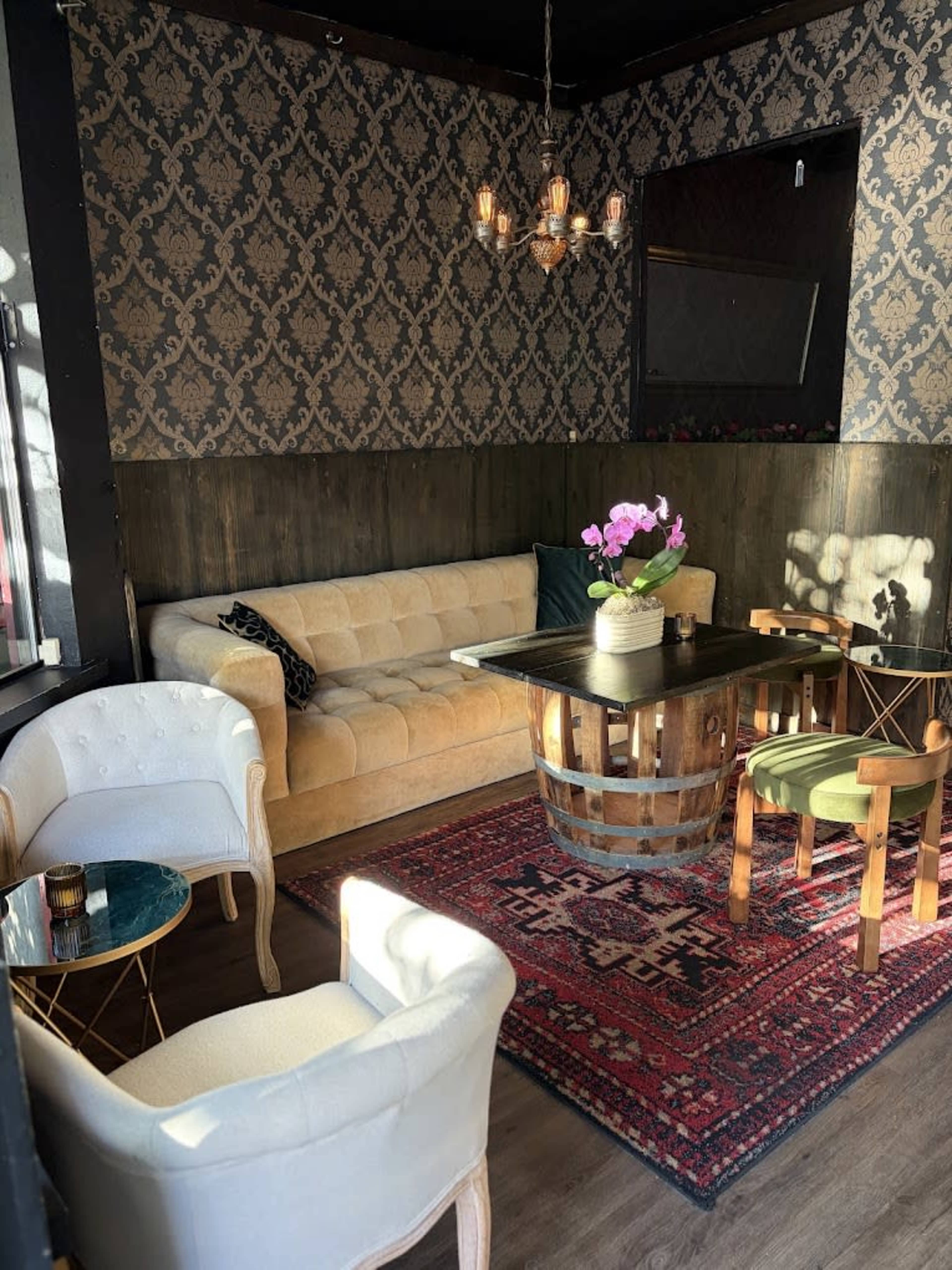 The image depicts a cozy seating area with a velvet sofa, wooden barrel table, and various chairs arranged around a patterned rug.