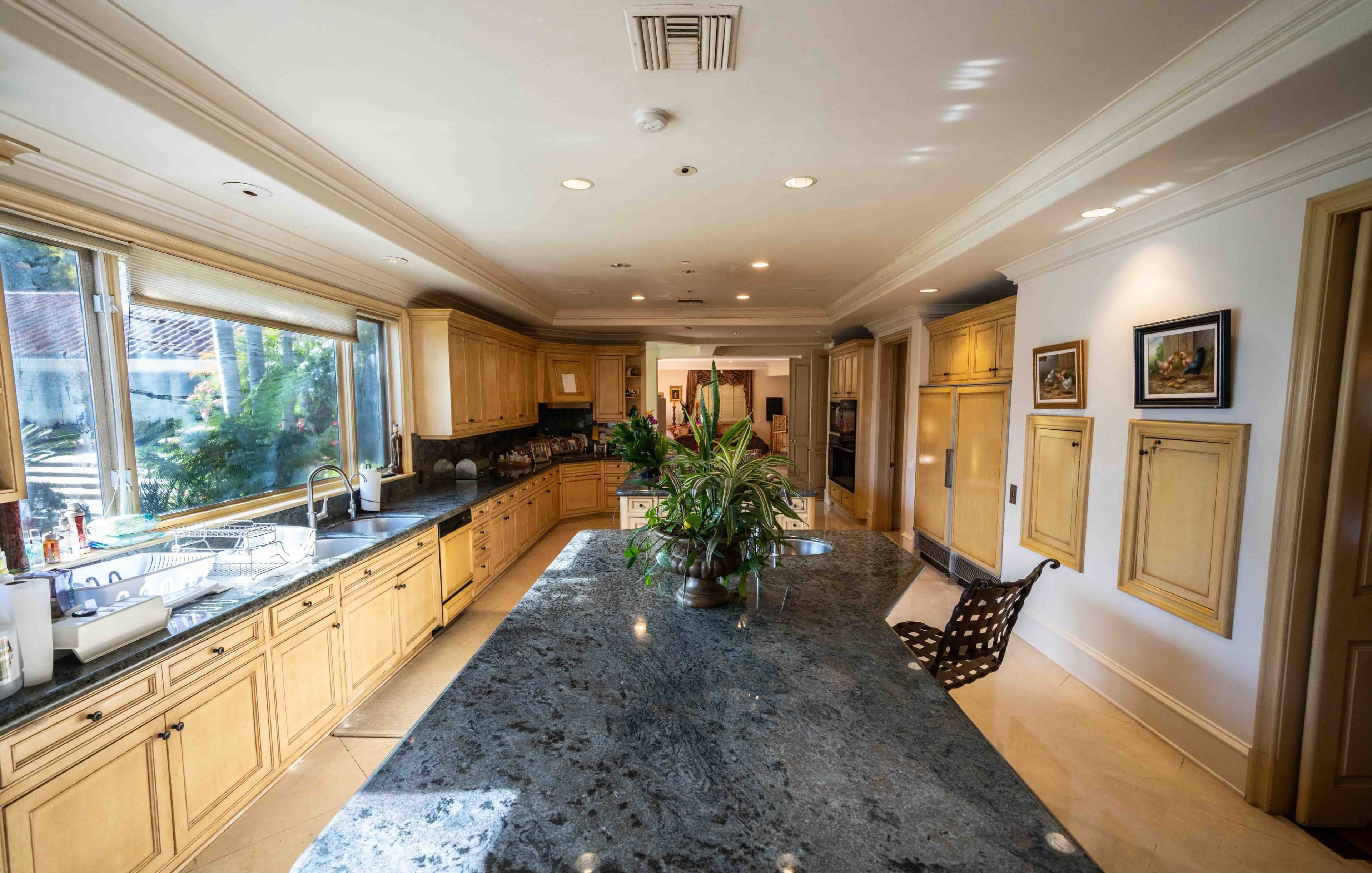 The image shows a spacious kitchen featuring wooden cabinetry, a large granite island, and ample natural light from large windows.