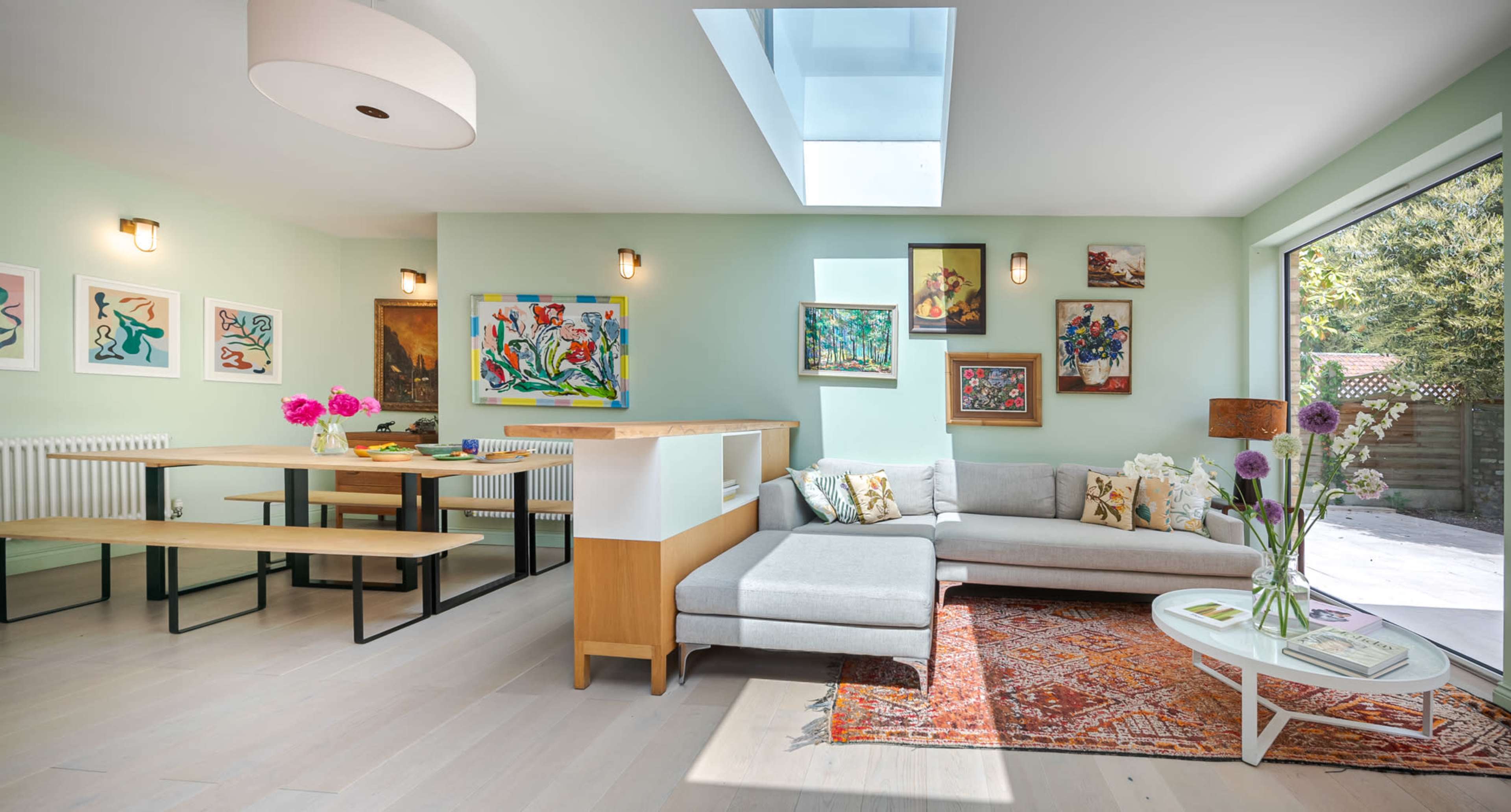 A modern living area with a light green wall, a sectional sofa, a coffee table, a dining space with benches, and several artworks displayed on the walls.