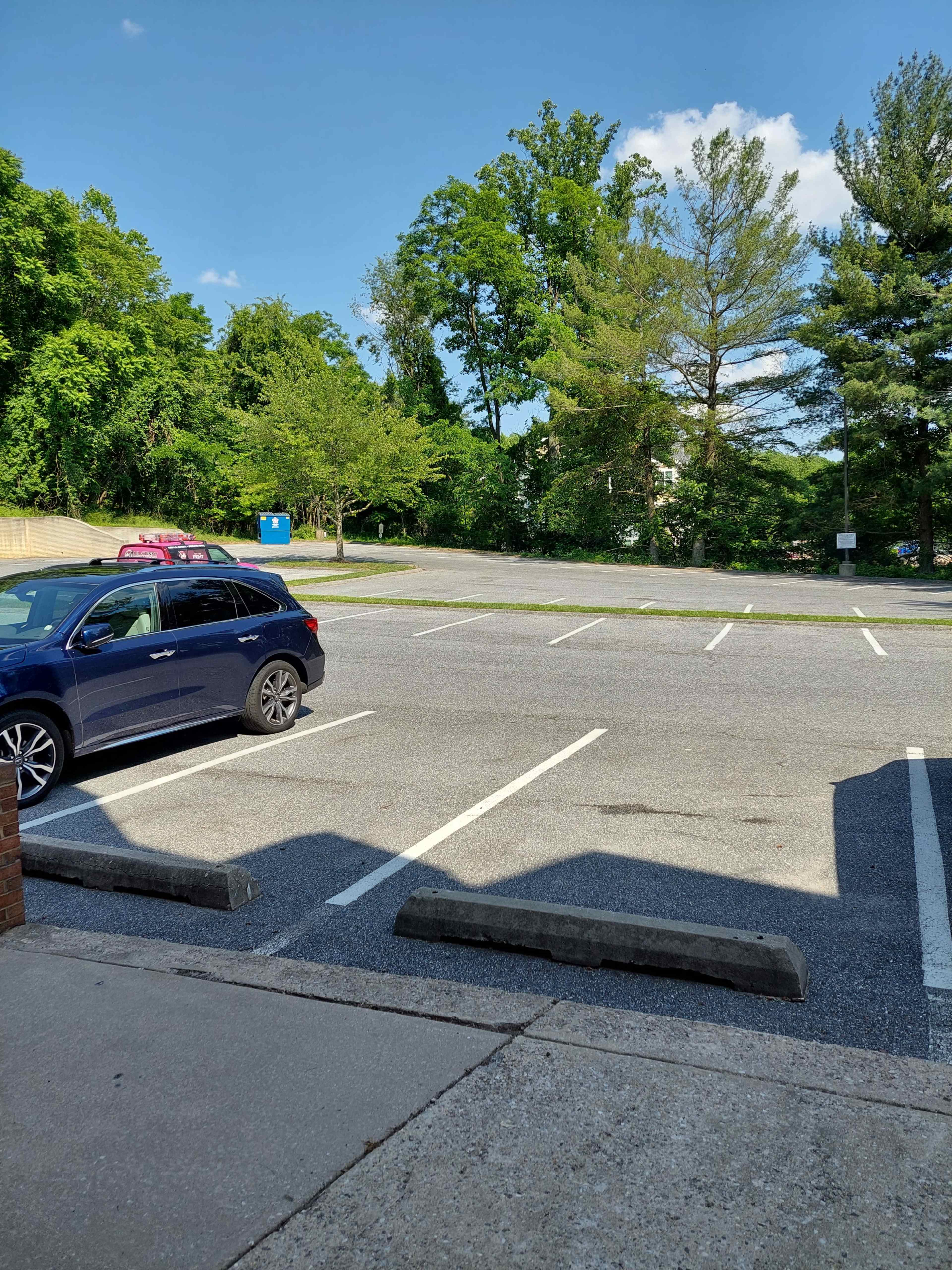 A blue car is parked in a nearly empty parking lot surrounded by trees and a clear sky.