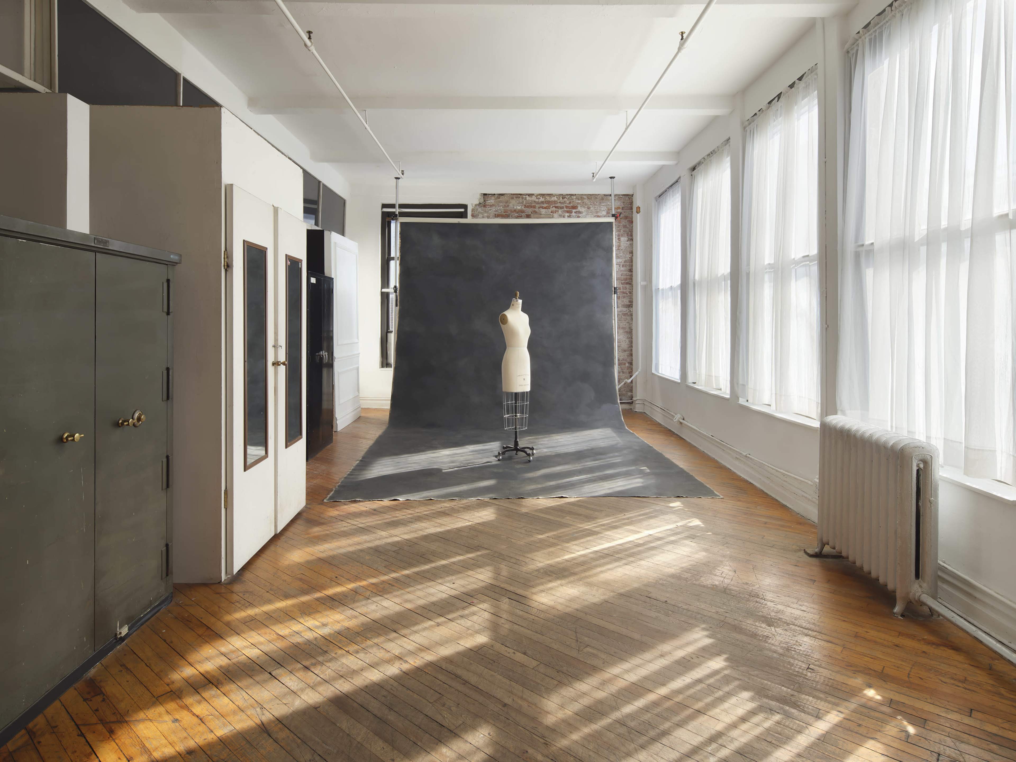 The image shows a spacious studio with a large black backdrop on the floor, a dress form on a stand in front of it, and windows allowing natural light to illuminate the wooden floor.