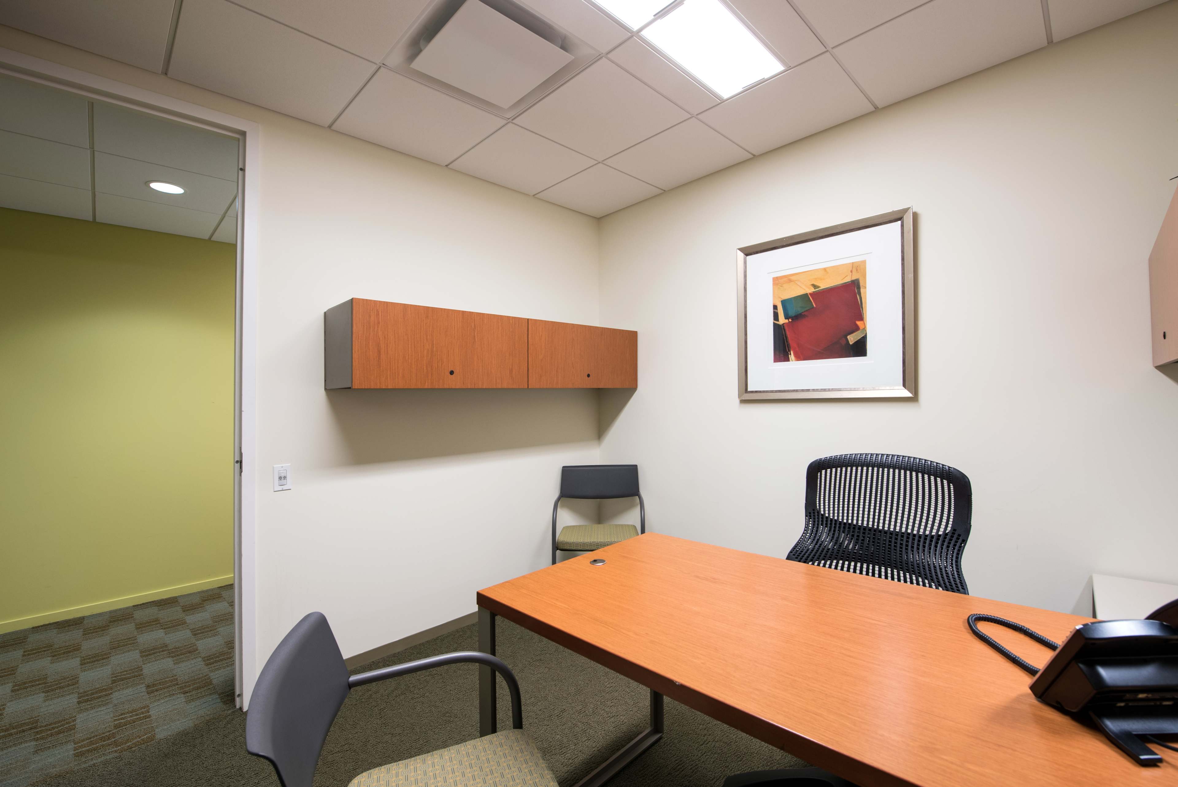 An office with a wooden desk, two chairs, a wall-mounted shelf, and a framed artwork is shown, featuring neutral-colored walls and carpeted flooring.