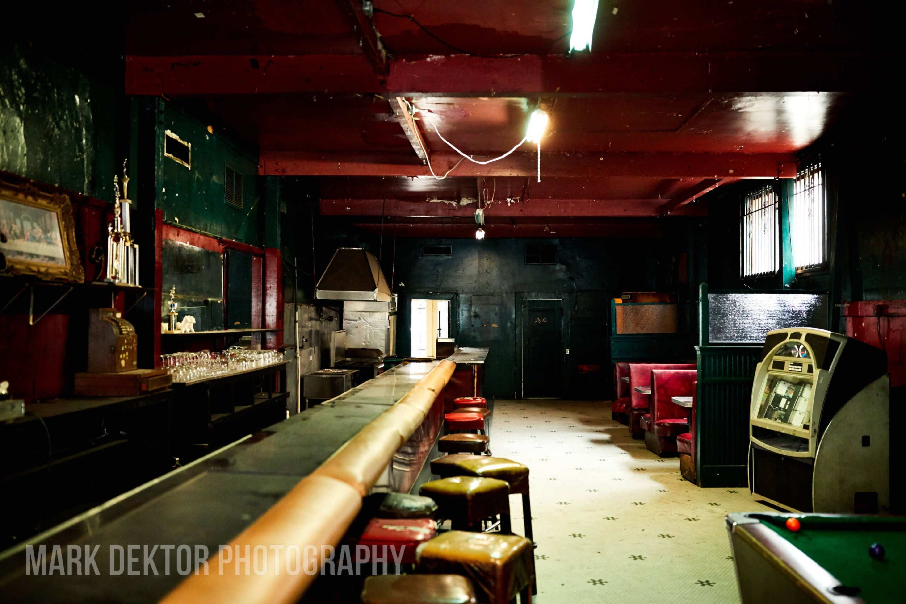 The image shows a dimly lit, empty bar with a long counter, stools, and a pool table in a quiet, untidy interior.