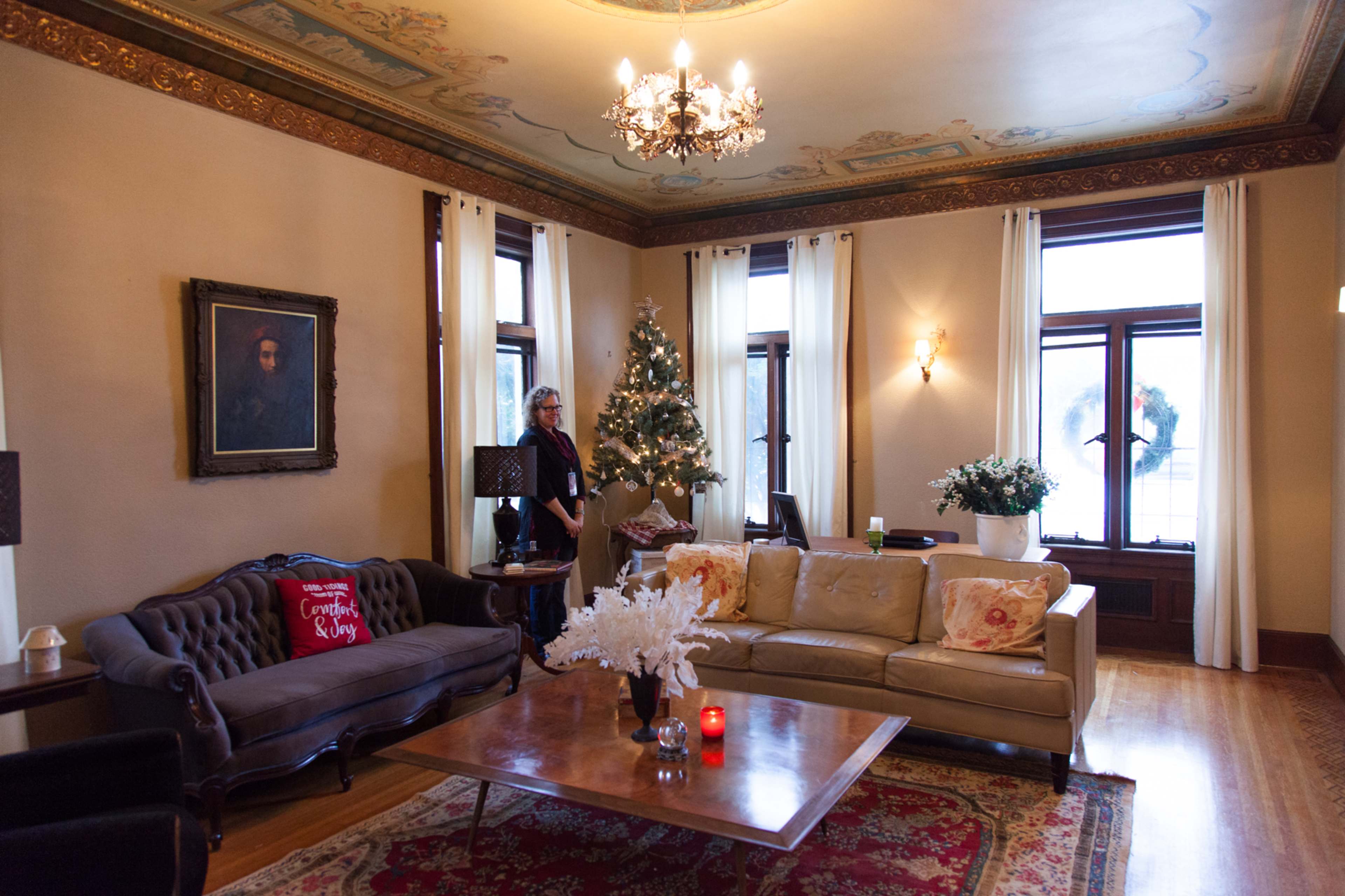 A cozy living room features a decorated Christmas tree, elegant furniture, and a large window adorned with sheer curtains.