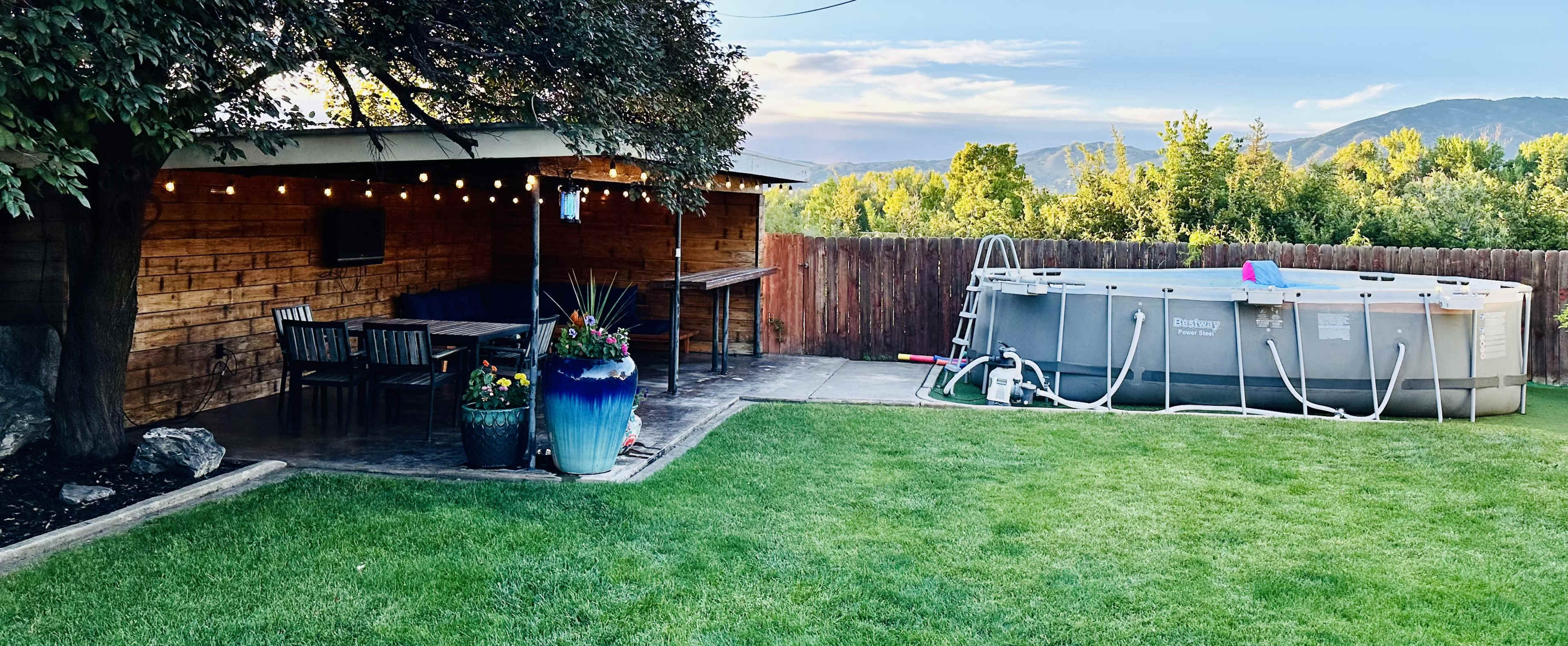 The image shows a landscaped backyard with a wooden patio, a dining area, and a round above-ground swimming pool.