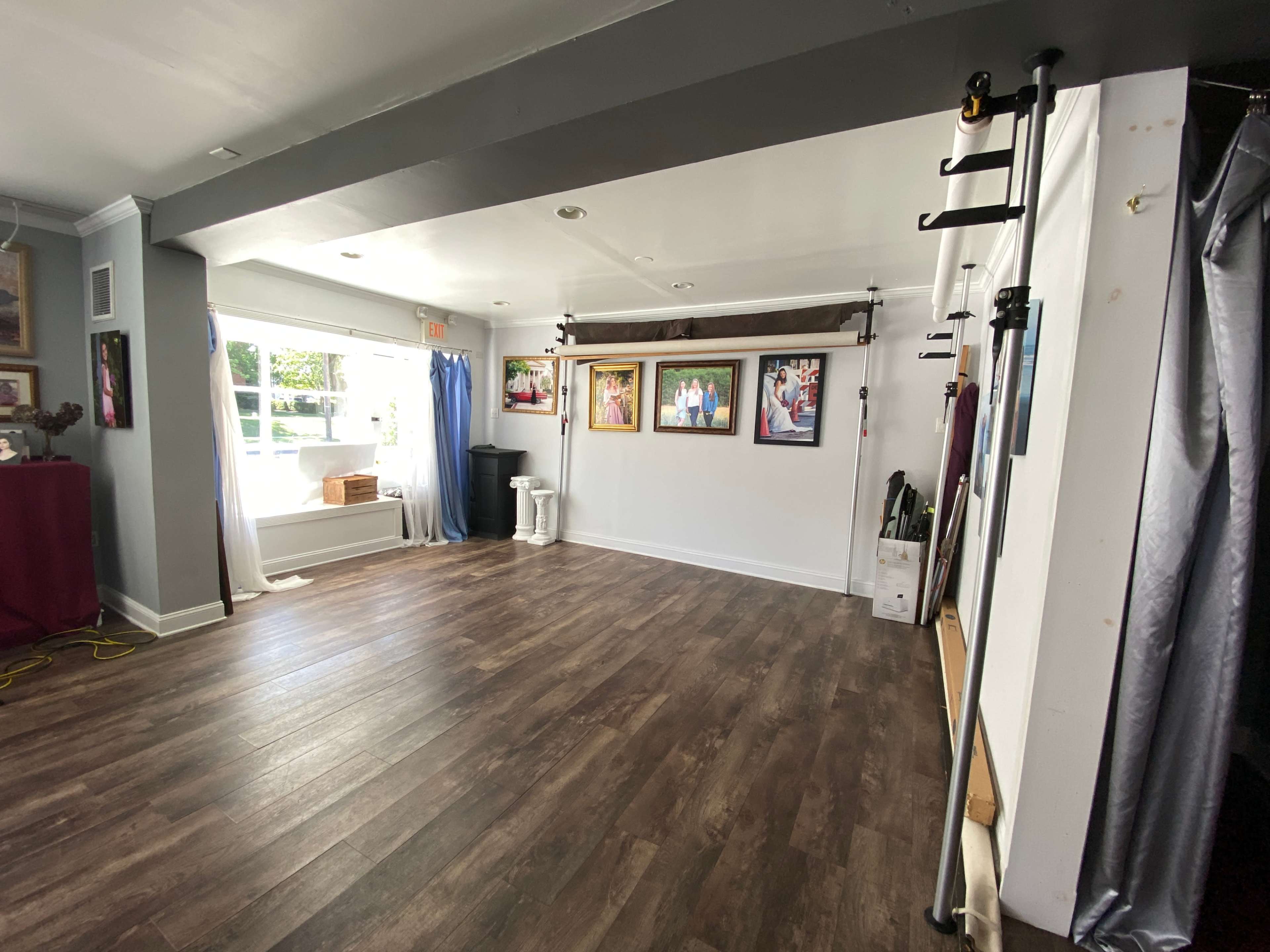 The image shows a spacious interior of a room with wooden flooring, framed photographs on the walls, and a large window that lets in natural light.
