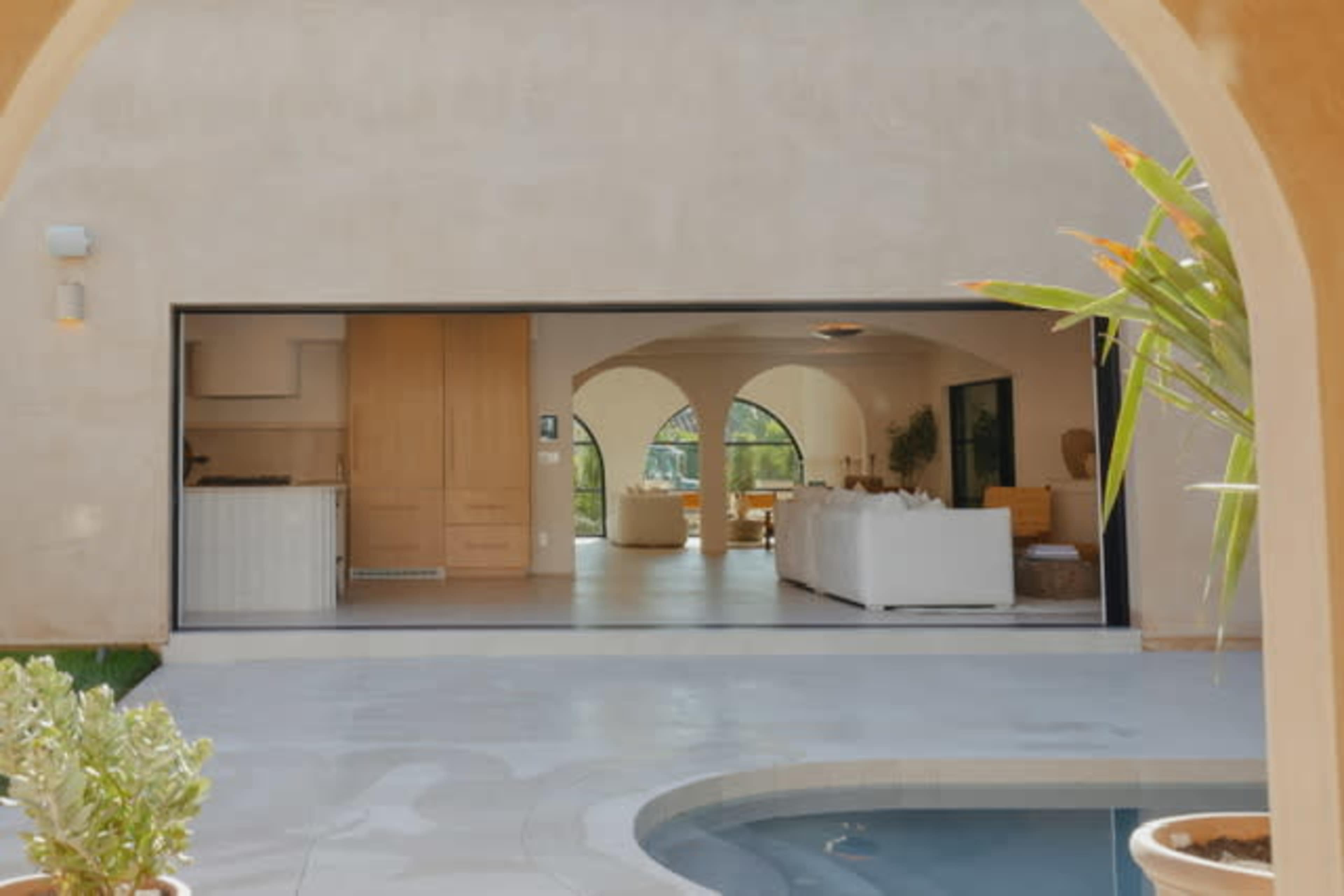 The image shows a modern, open-concept living area with large arches and sliding glass doors opening to a patio and swimming pool.