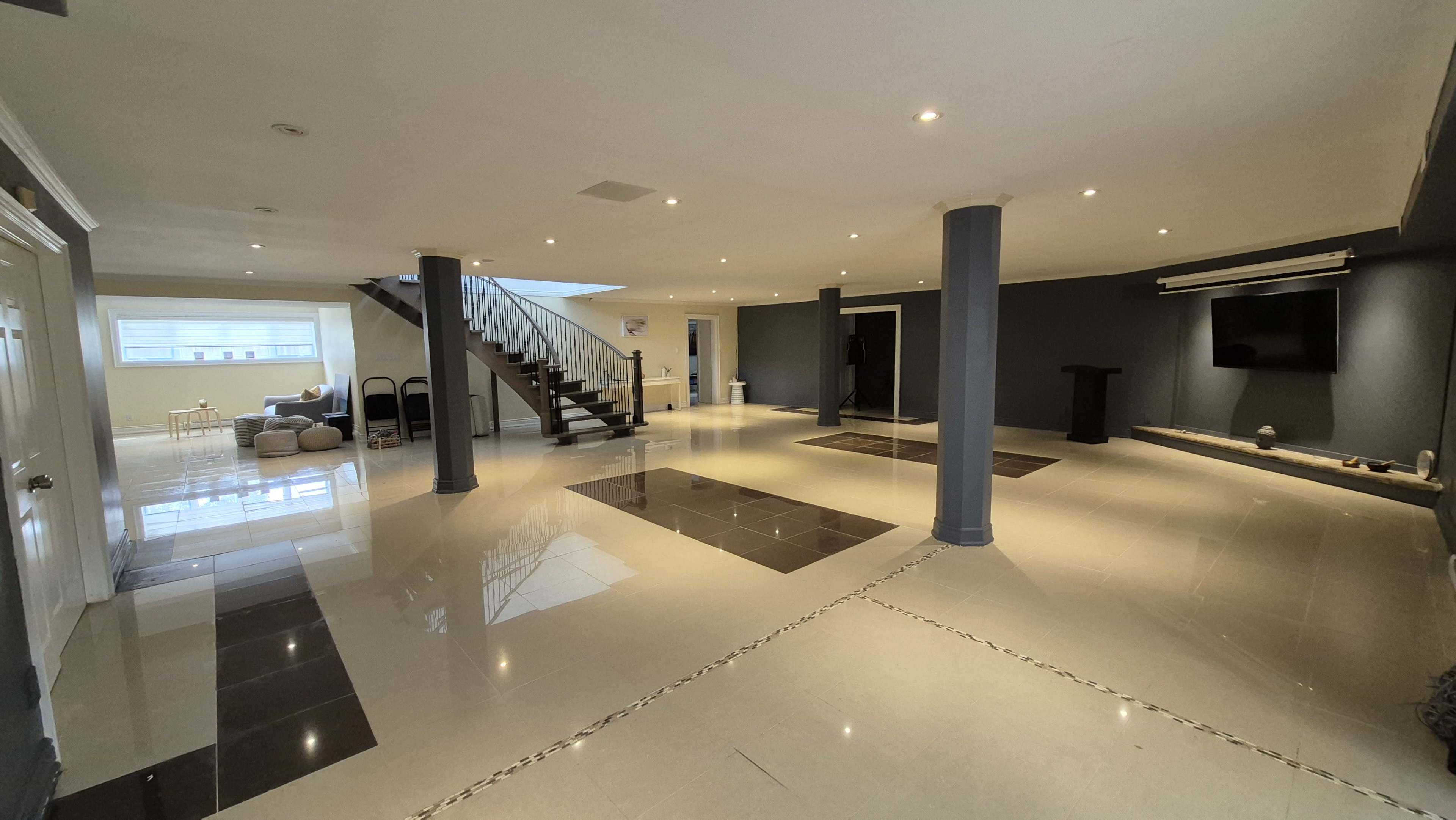 The image shows a spacious, well-lit room with polished tile flooring and a staircase in the background.
