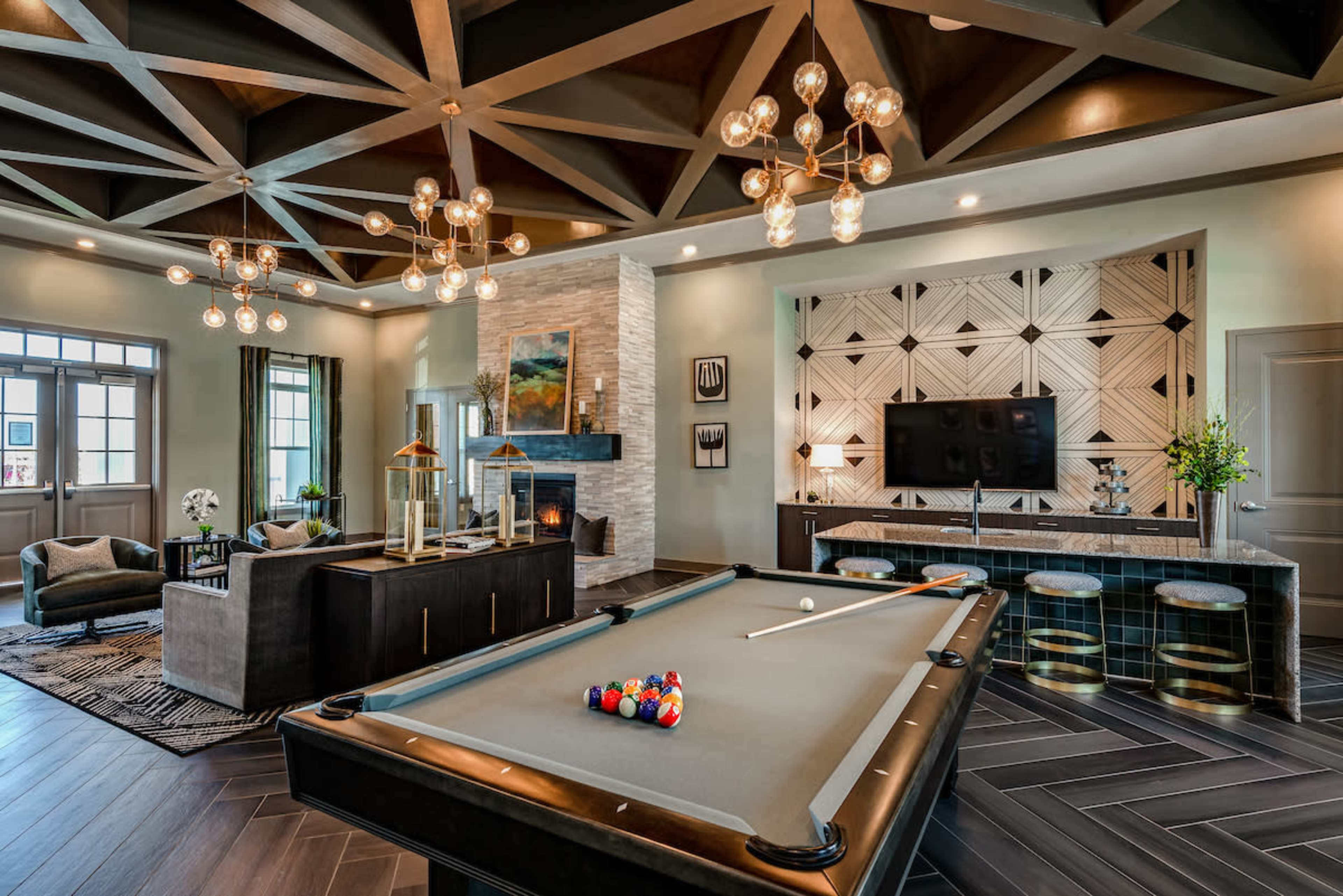 The image shows a modern game room featuring a billiard table, a stone fireplace, a television, and stylish lighting fixtures.
