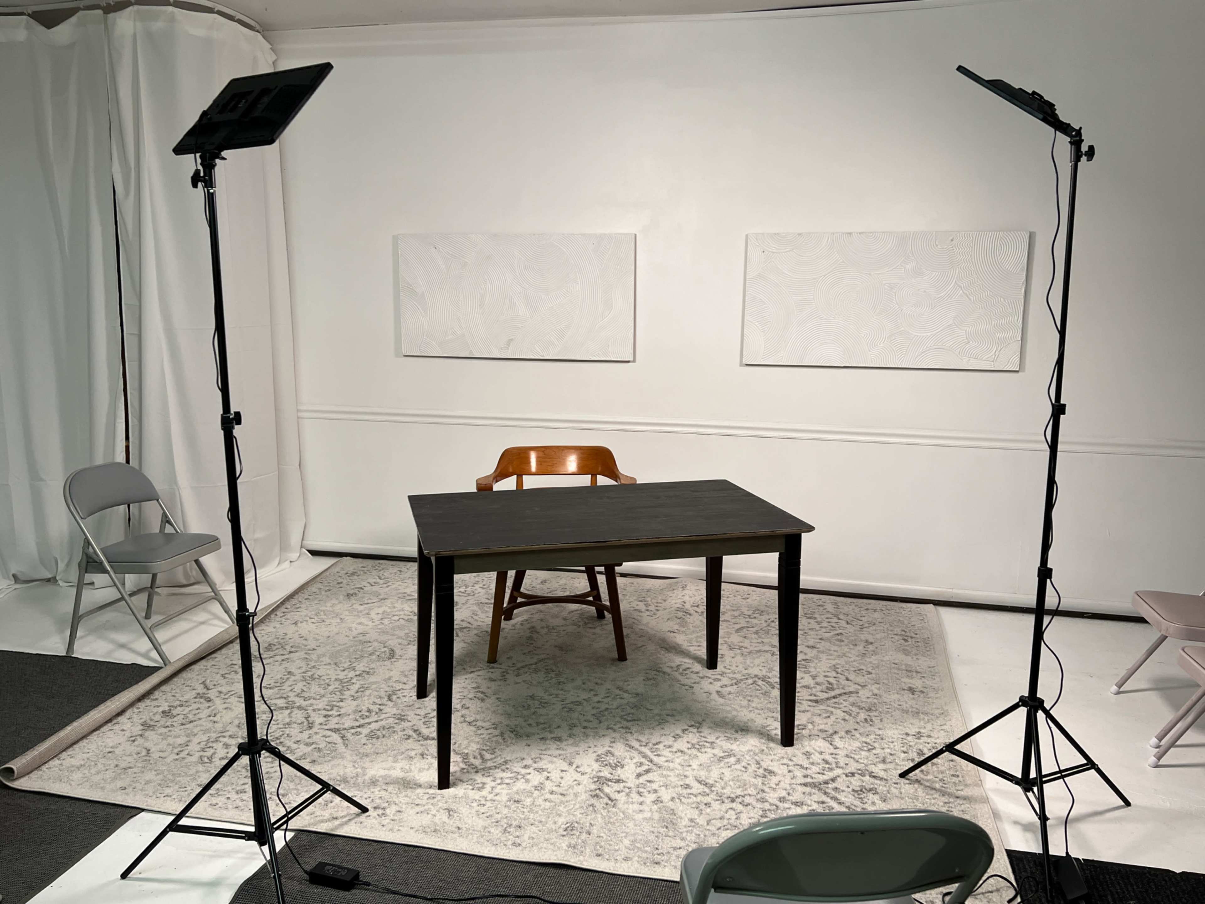 A minimalistic studio setup featuring a wooden table and a chair on a rug, with two lights and white wall panels in the background.